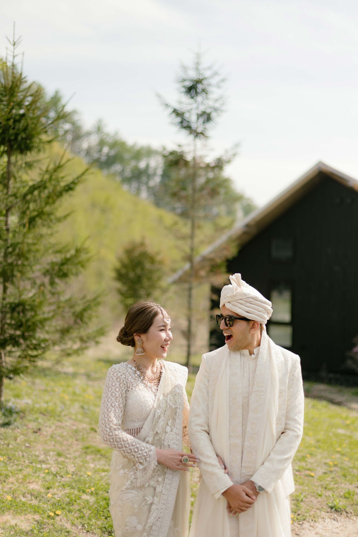 Indian wedding couple in white traditional attire laugh together in scenic outdoor countryside setting.