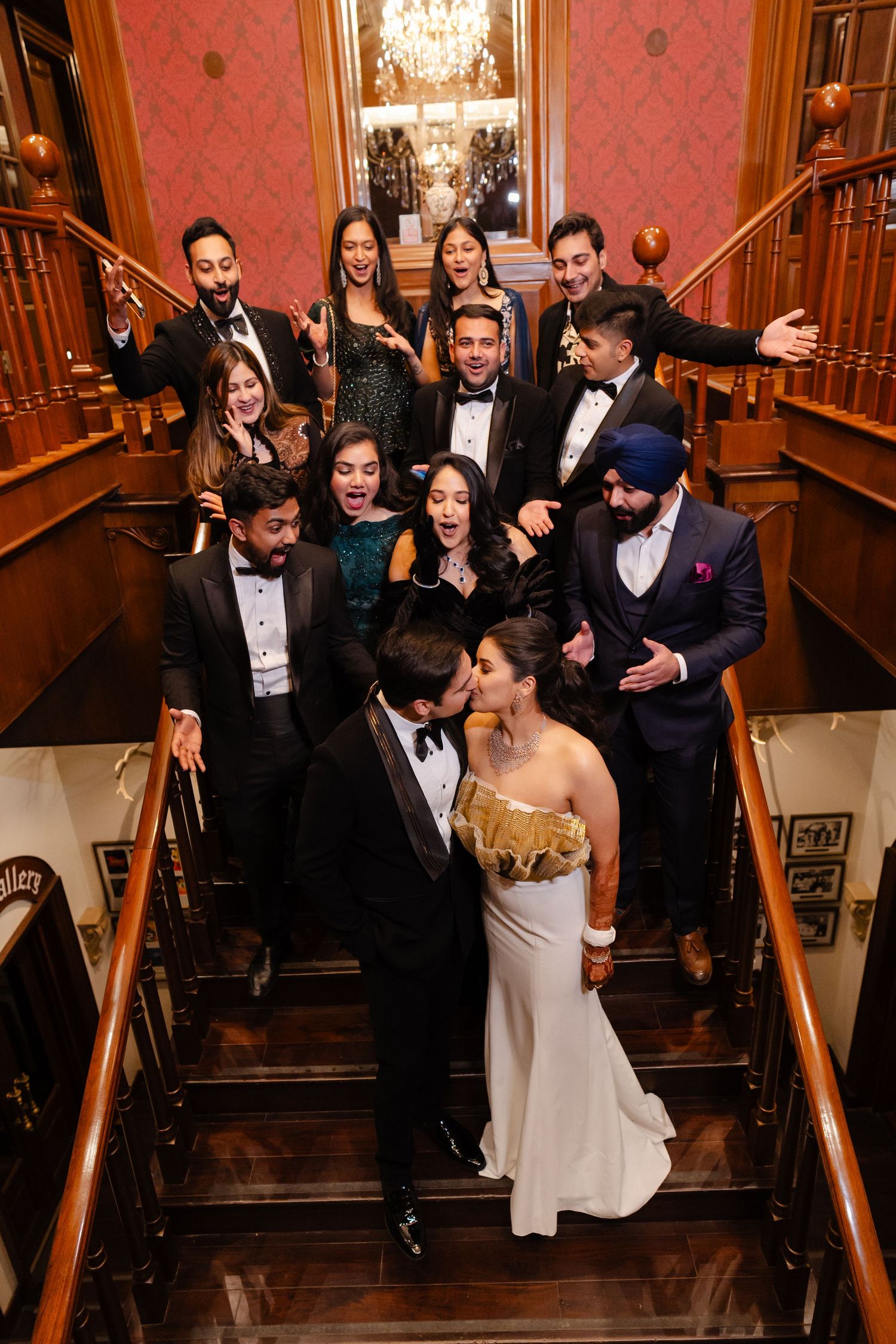 Wedding party celebrating on grand staircase, bride and groom kissing surrounded by cheering guests in formal attire.