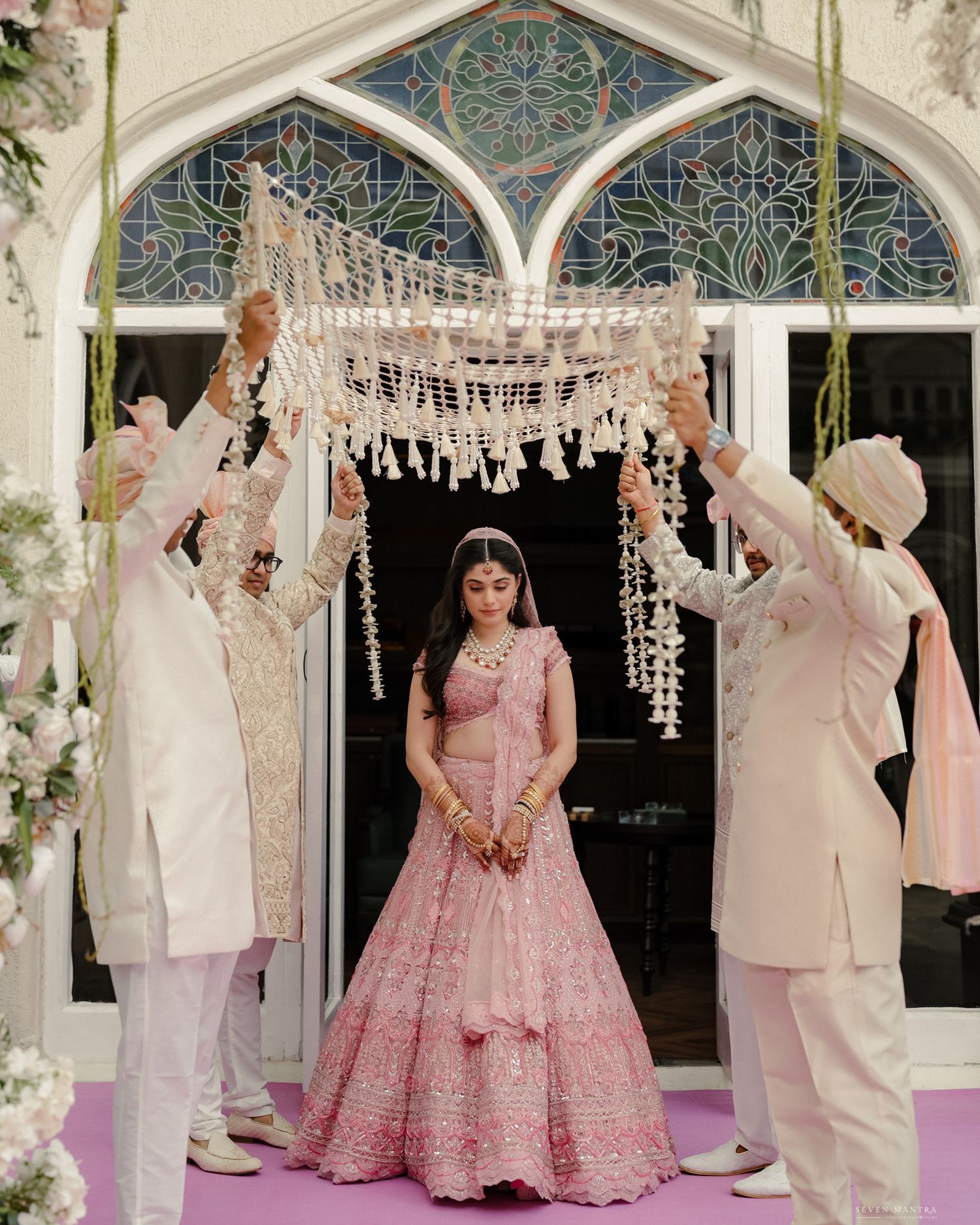 Indian bride in pink lehenga making grand entrance under floral chandni at wedding ceremony.