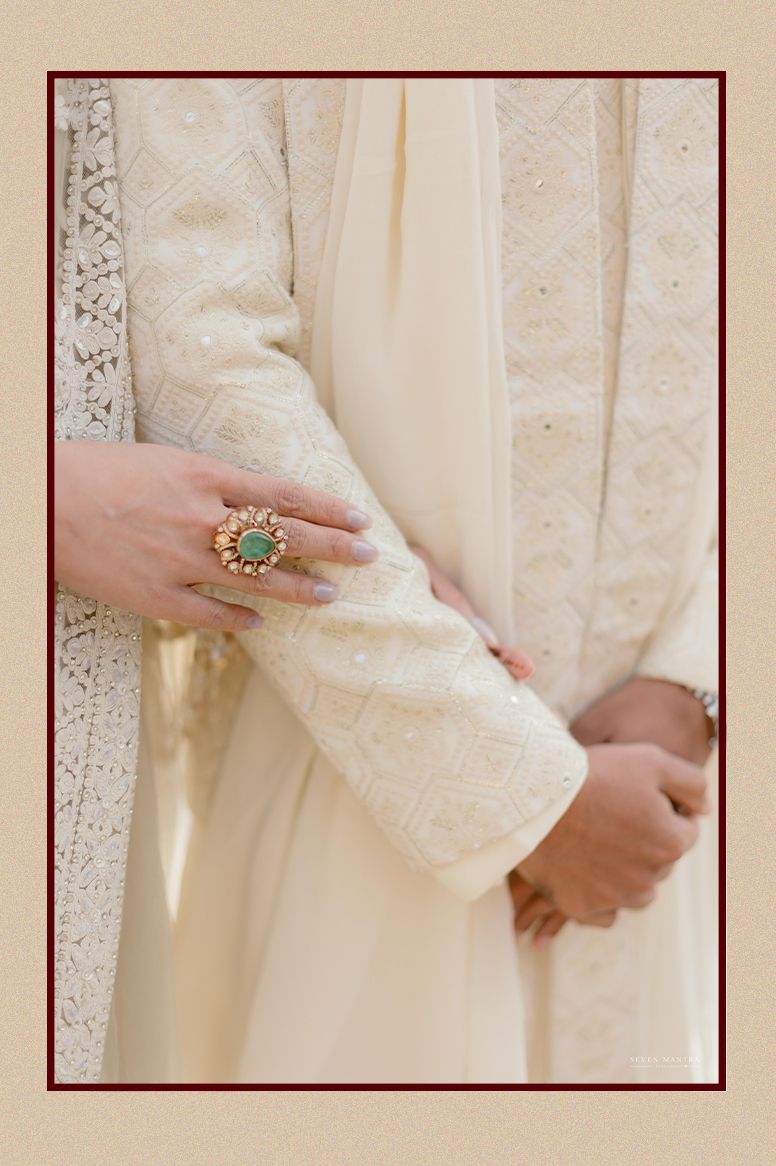 Close-up of bride and groom hands with gold emerald ring against ivory embroidered sherwani fabric.