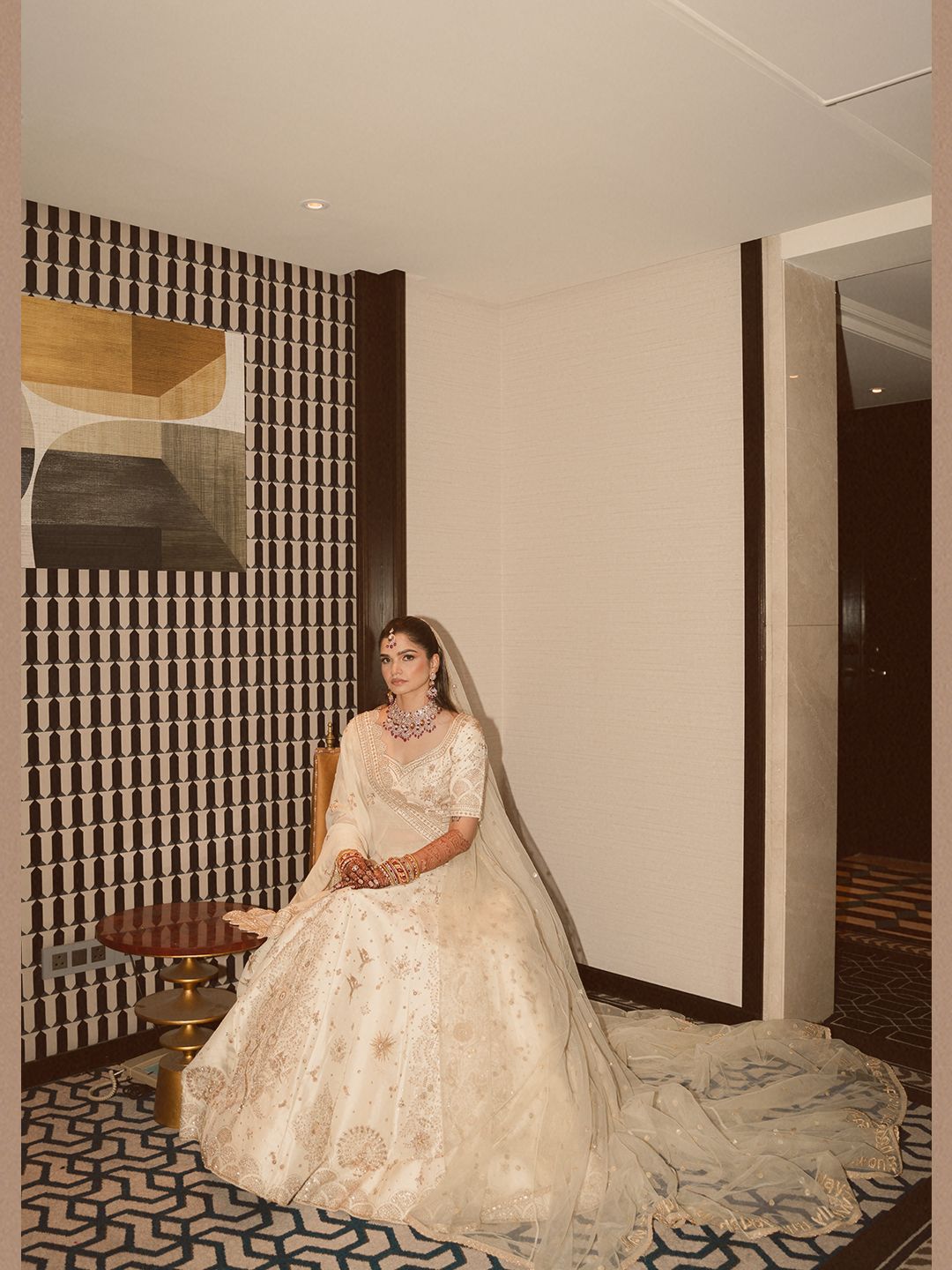 Bride in ivory embroidered lehenga with veil seated elegantly in a luxury hotel corridor.