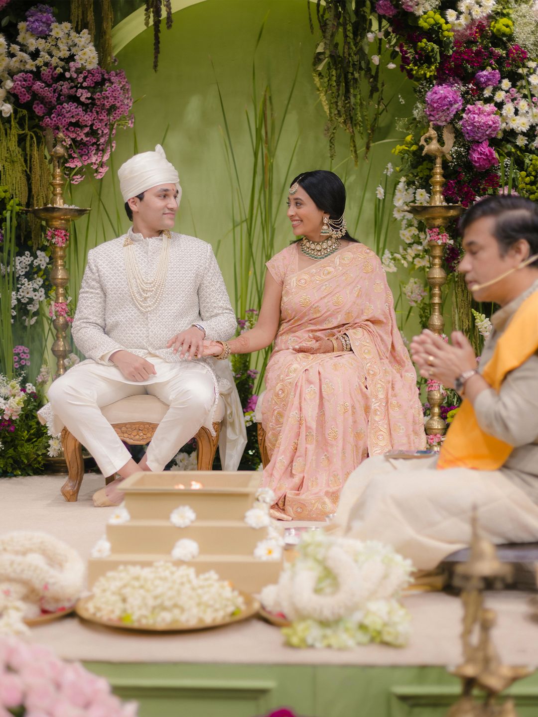 Indian bride in pink lehenga and groom in white sherwani during Hindu wedding ceremony with priest.