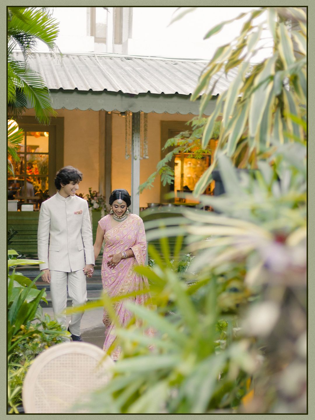 Couple walks hand in hand through tropical garden pathway near elegant venue entrance.