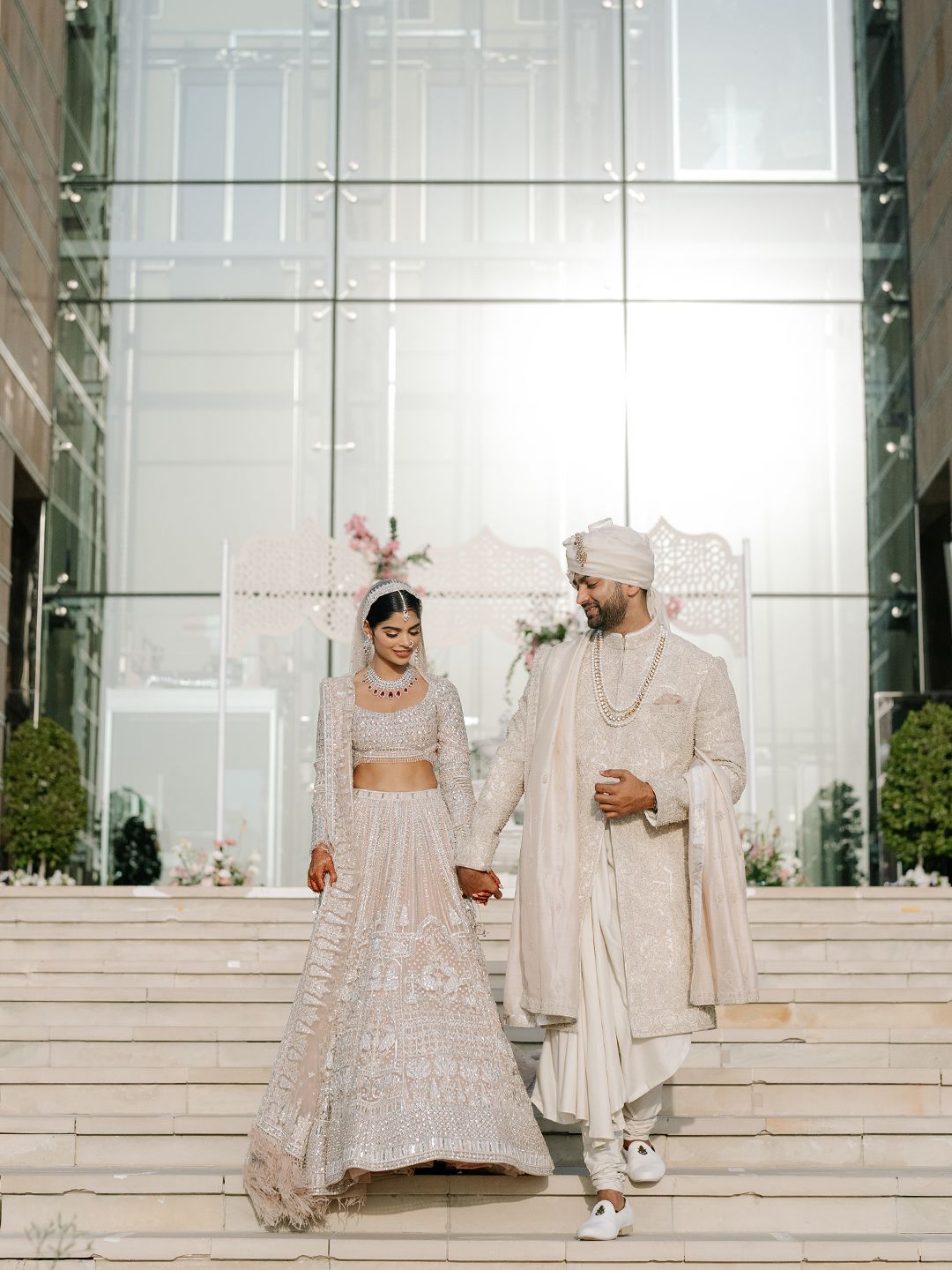 Indian bride and groom in ivory embroidered outfits hold hands descending steps at elegant glass venue.