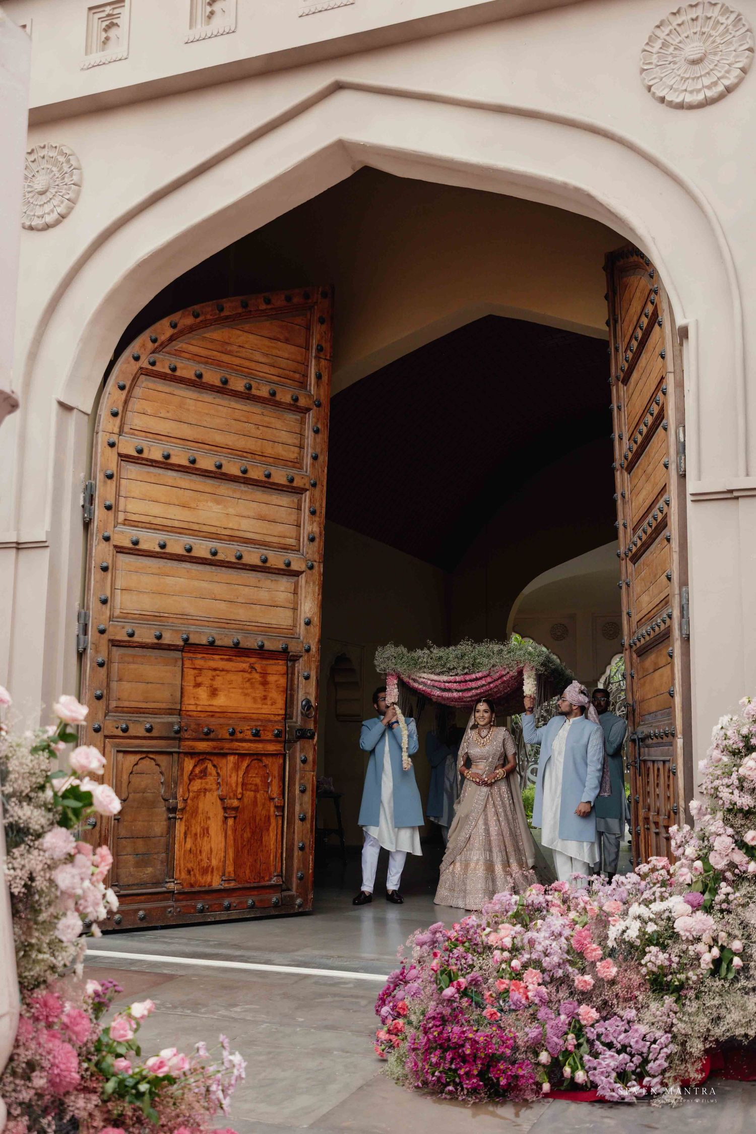 Bride making grand entrance through ornate wooden doors adorned with pink floral arrangements.