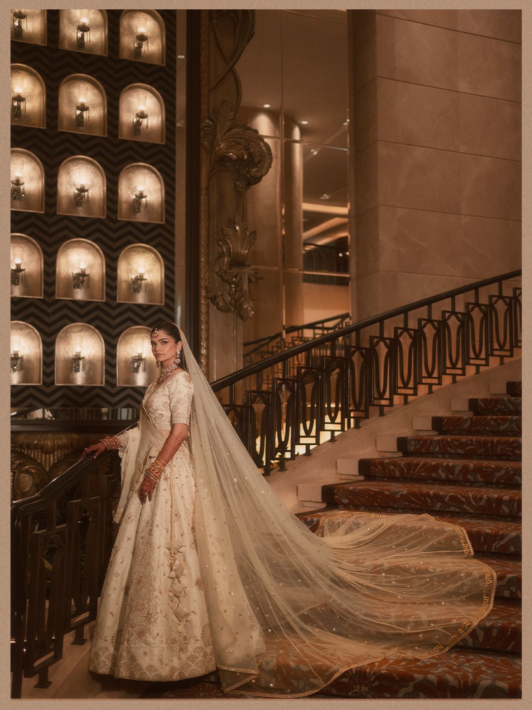 Bride in ivory embroidered lehenga with long veil posing on grand staircase in luxurious hotel lobby.