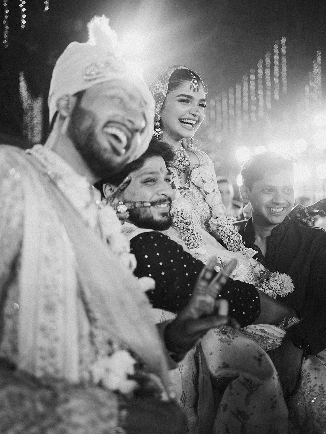 Joyful black and white photo of bride and groom laughing during Indian wedding ceremony with guests.