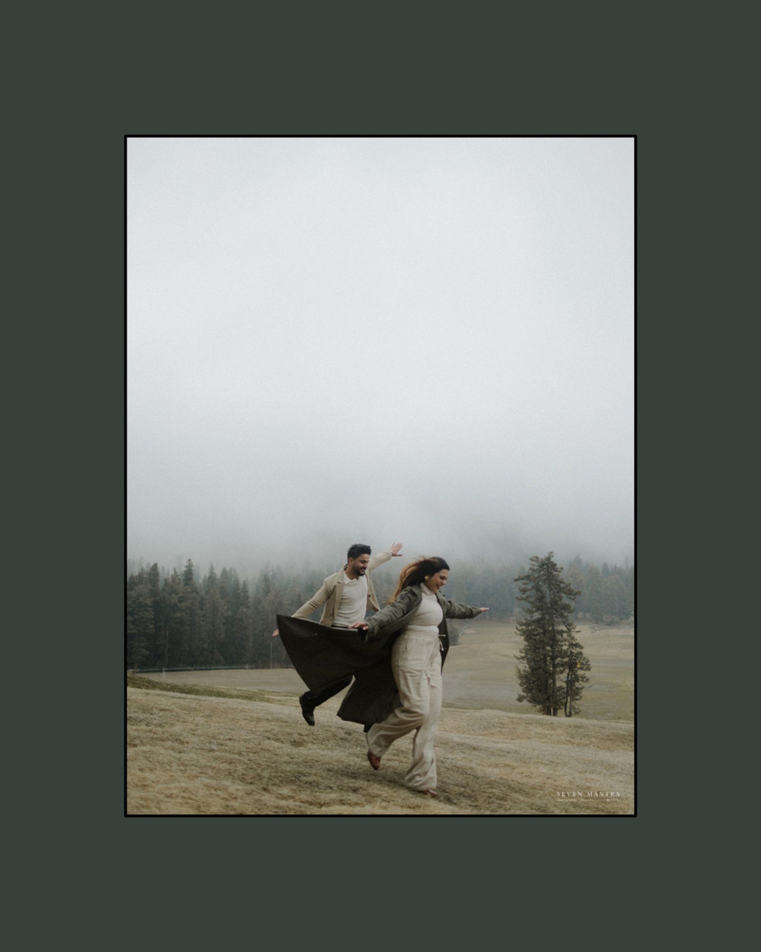 Indian couple playfully running through a misty foggy meadow surrounded by pine trees.