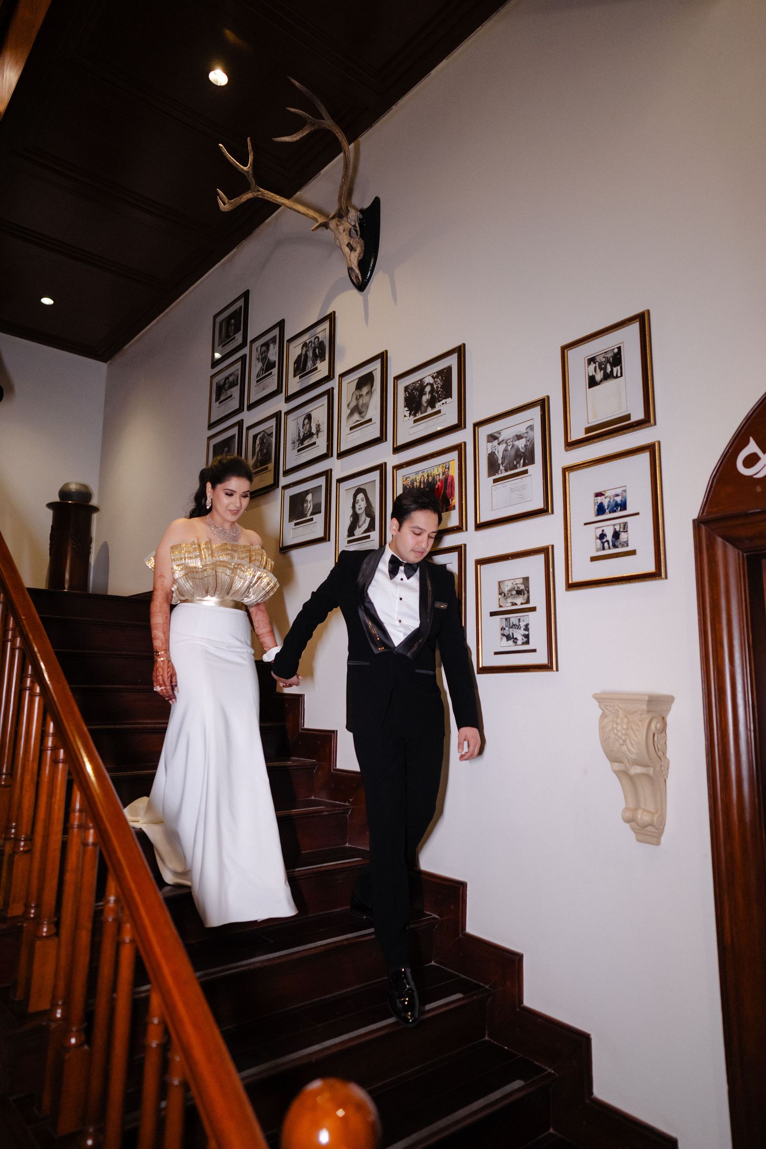 Elegant couple descending wooden staircase, bride in white gown with gold top, groom in black tuxedo.