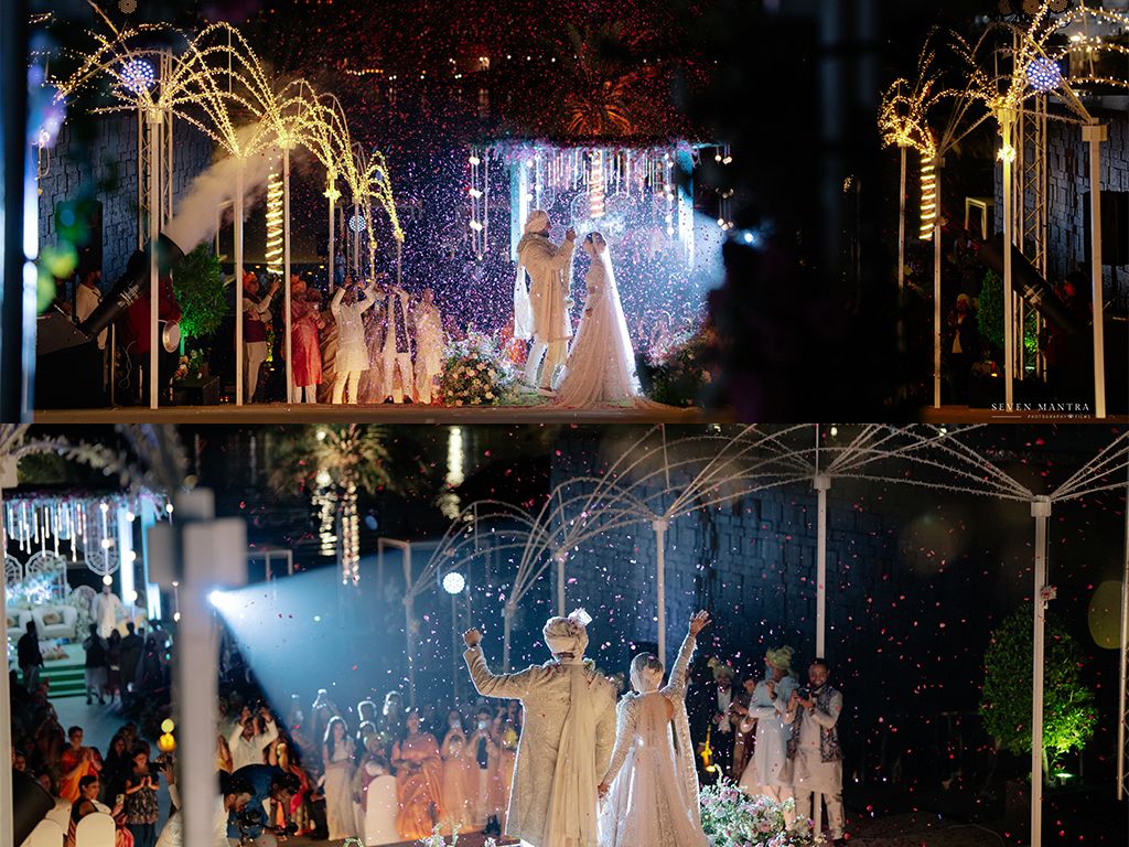 Indian wedding couple celebrates on stage with sparkling pyrotechnics and dancing guests at grand reception.