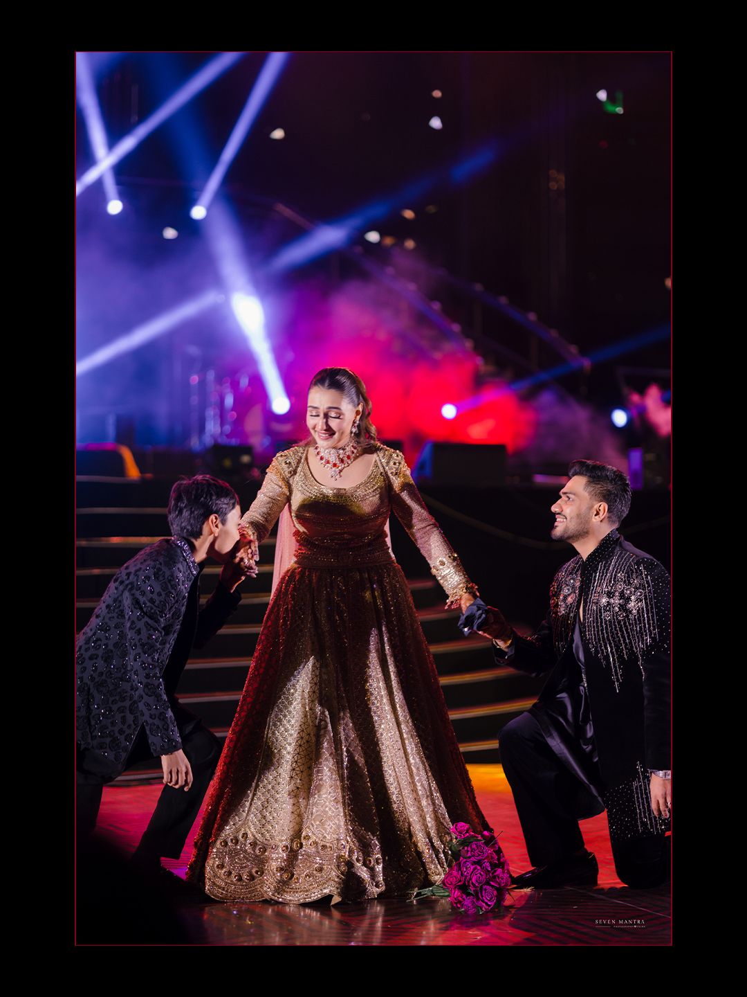 Bride in golden brown lehenga laughing joyfully on stage flanked by two men under colorful event lighting.