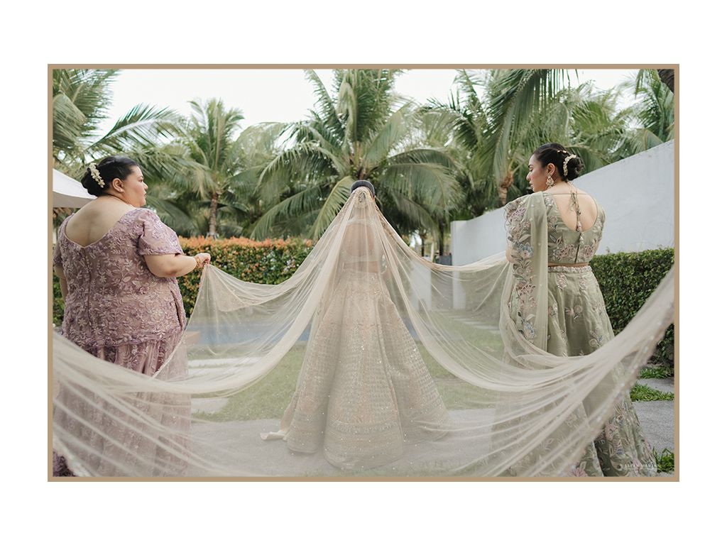 Two women in pink sarees spread elegant long white bridal veil outdoors among tropical palm trees.