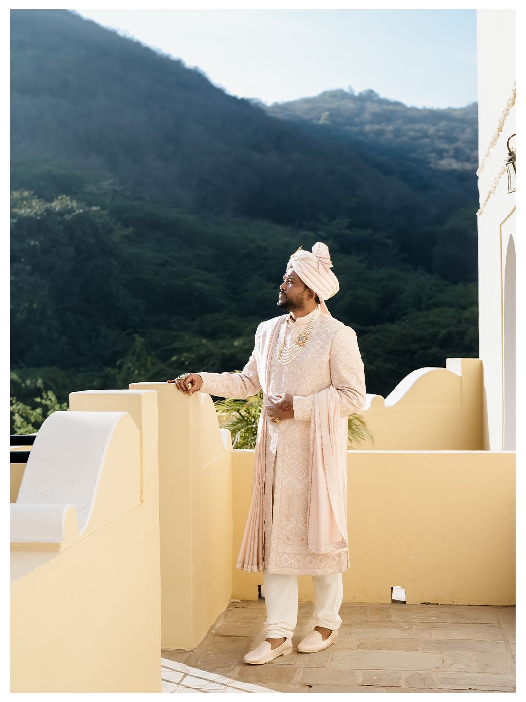 Groom in pink sherwani and turban standing on terrace with lush green mountains behind him.