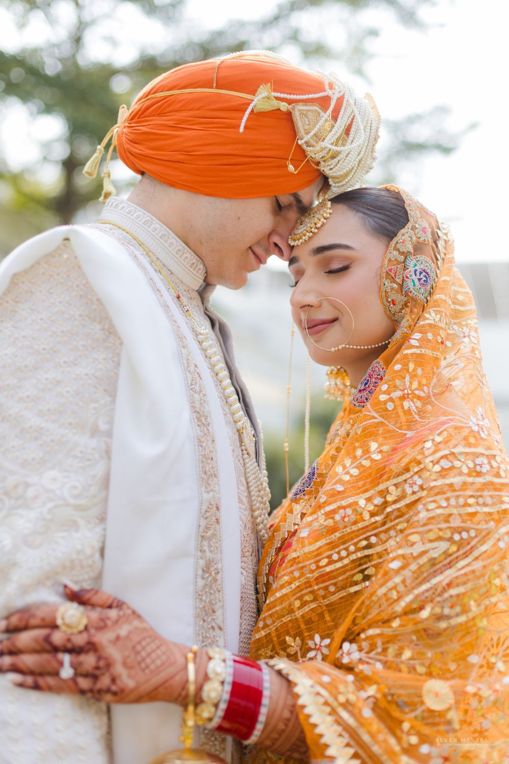Sikh bride and groom in orange and ivory wedding attire share an intimate forehead touch moment.