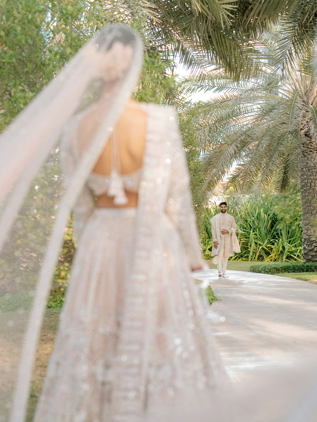 Groom walks toward bride in embroidered lehenga with flowing veil on palm-lined wedding venue pathway.