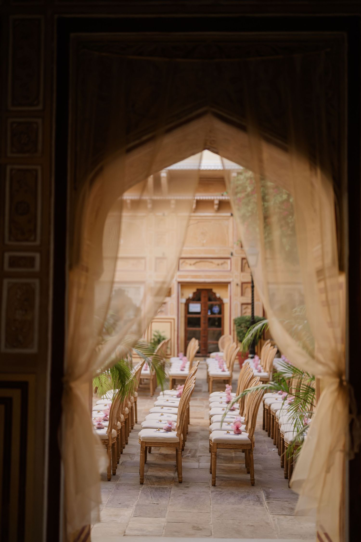 Elegant Indian wedding reception setup viewed through ornate Mughal arch with draped curtains and decorated tables.
