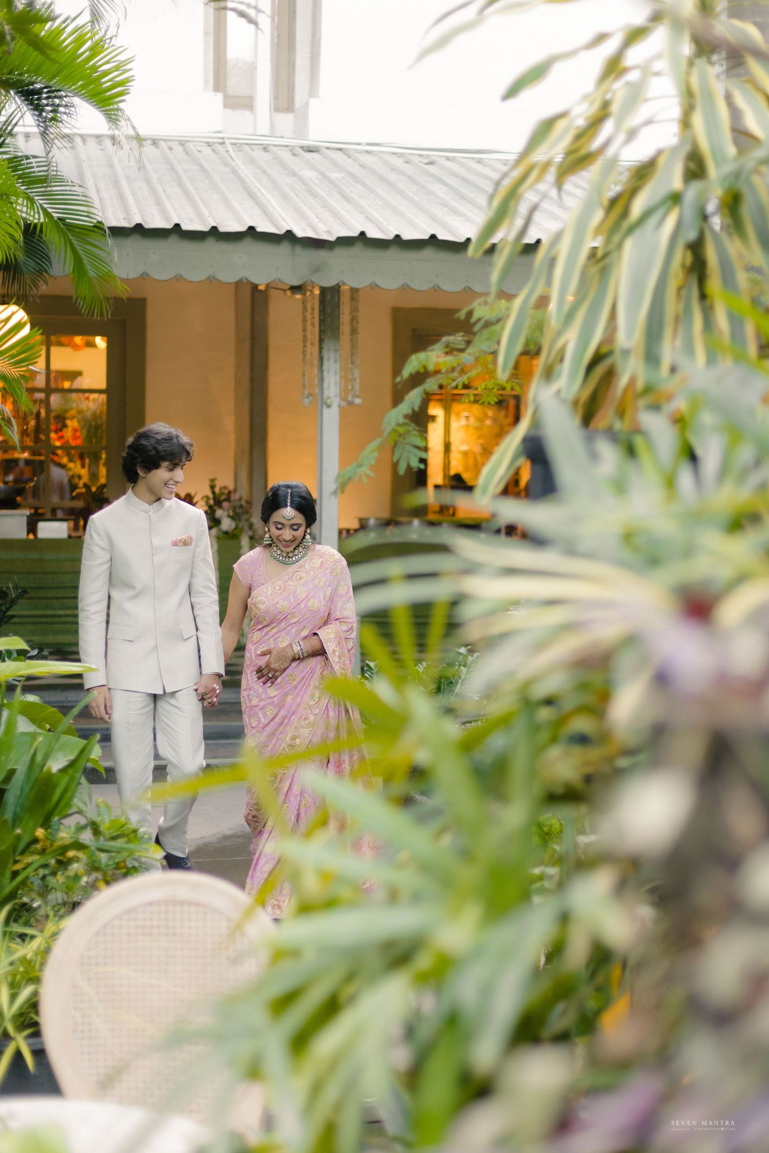 Indian couple in traditional wedding attire walking hand-in-hand through a lush garden courtyard.