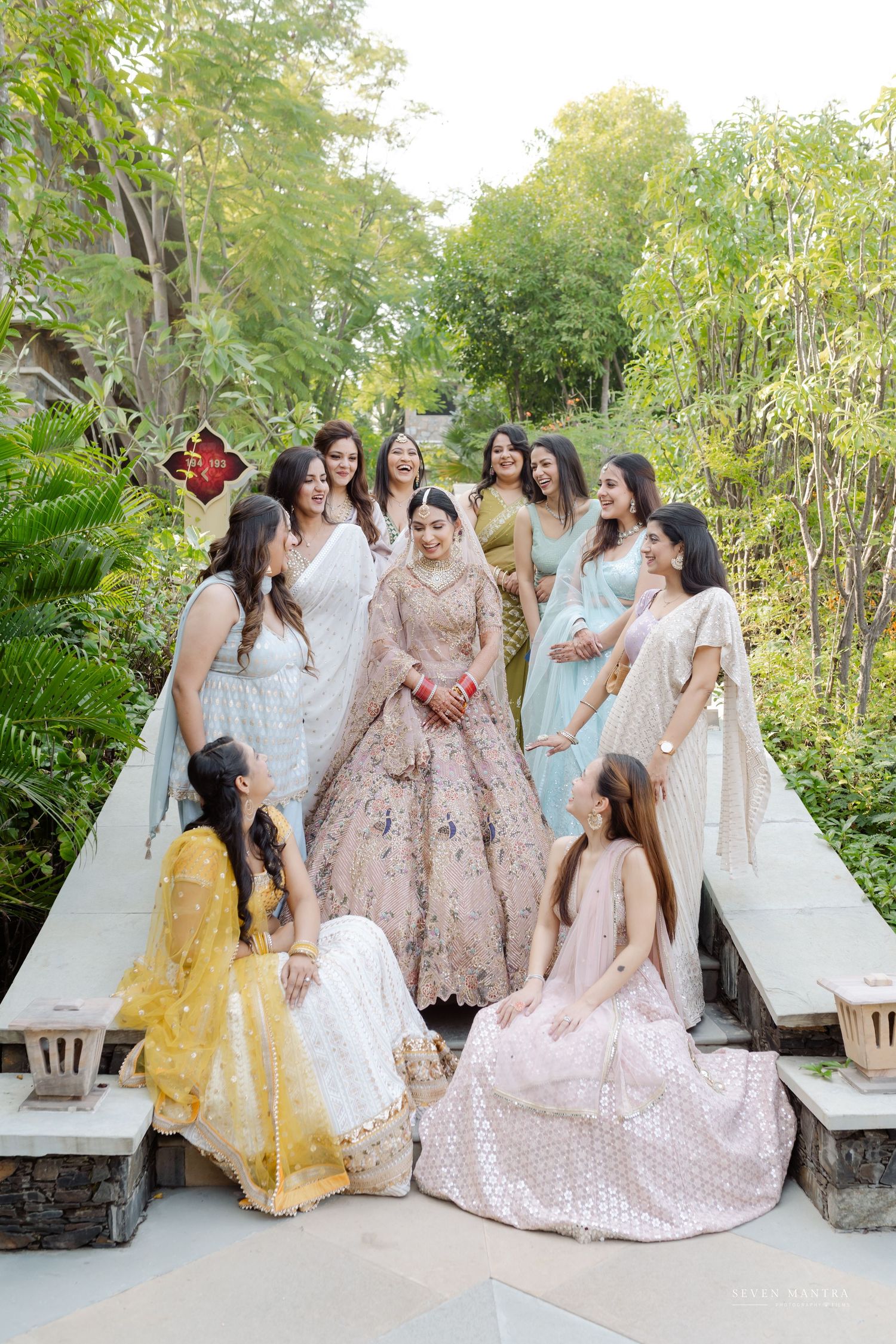 Indian bride in pink lehenga surrounded by bridesmaids in pastel sarees and lehengas in a lush garden setting.