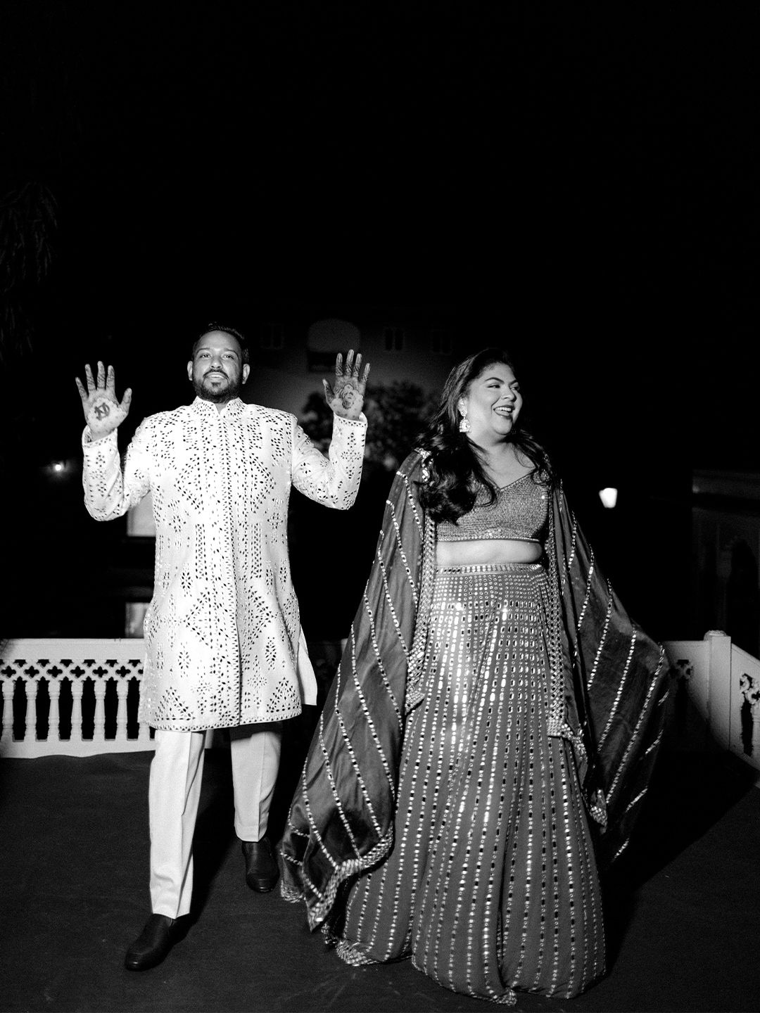 Couple in traditional Indian attire waving at their wedding celebration in black and white photography.