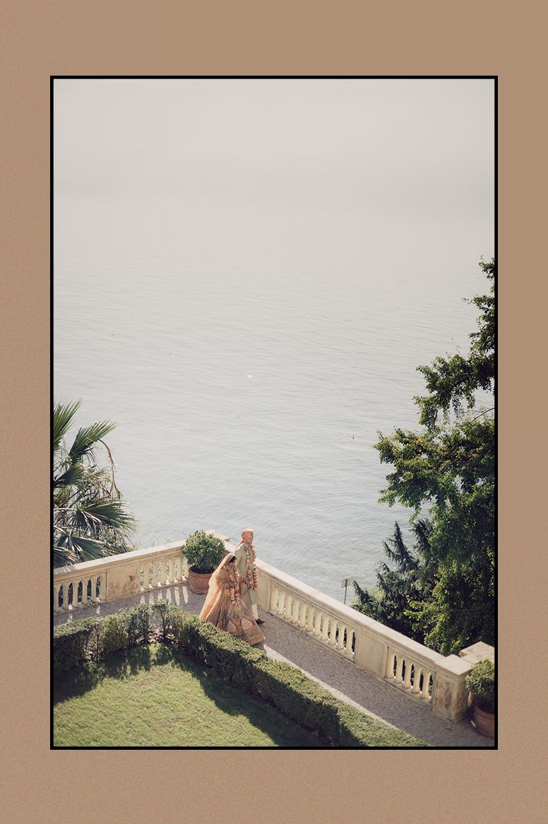 Indian bride in peach lehenga standing on terrace overlooking serene lake surrounded by lush greenery.