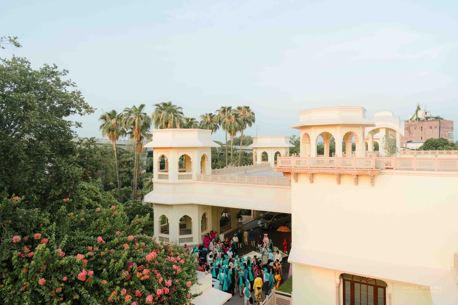 Aerial view of elegant Indian palace wedding venue with palm trees and colorful guests.