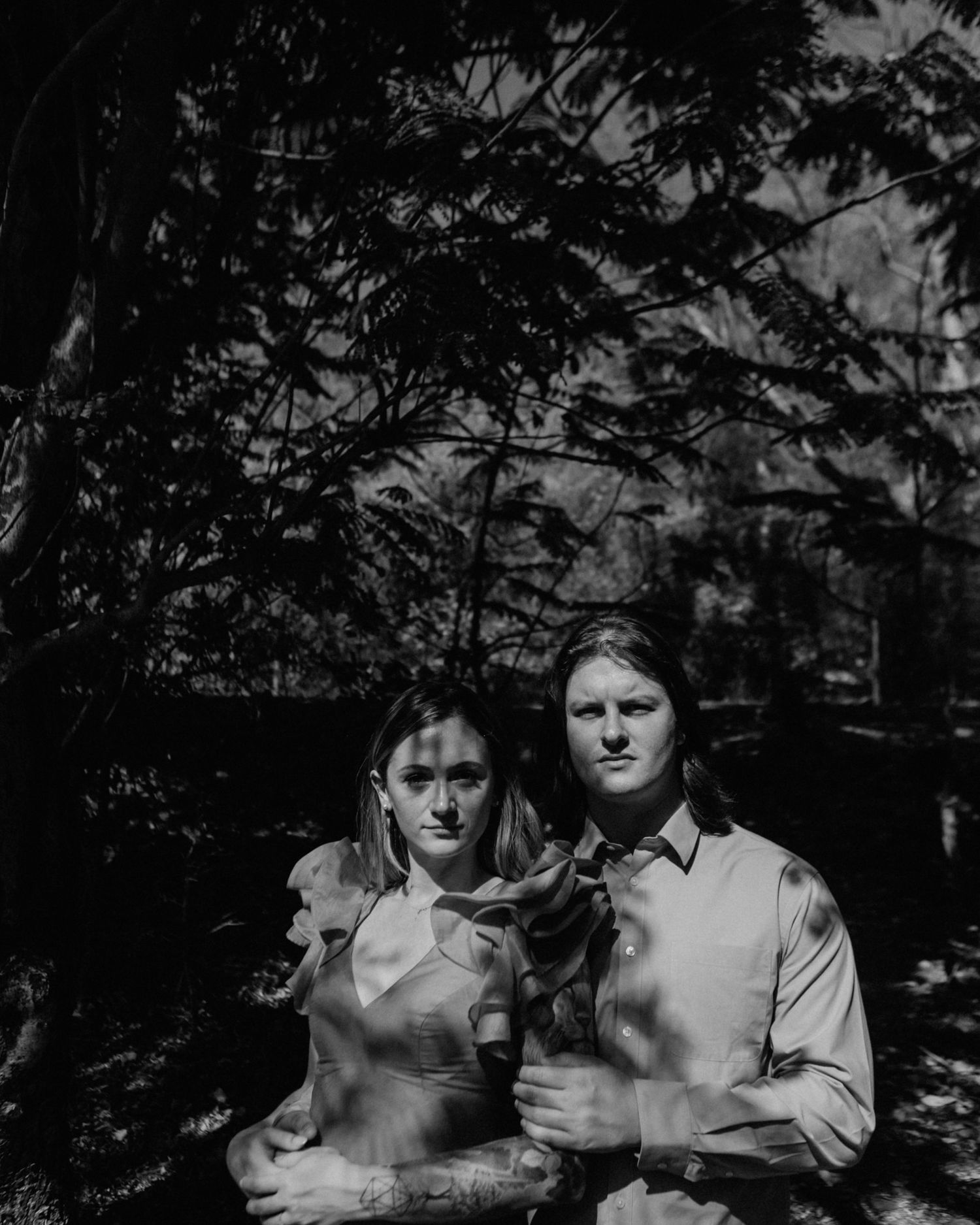 Black and white portrait of a couple standing close together under dark forest trees.
