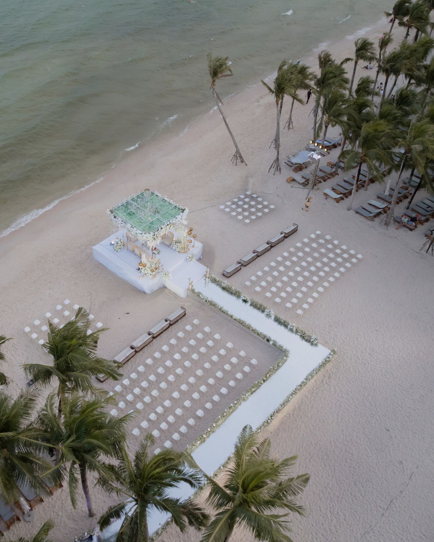 Aerial view of elegant beachfront wedding mandap setup with white chairs and palm trees.