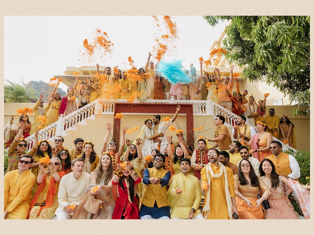 Large wedding party in yellow and orange attire throws colored powder at lively haldi celebration.