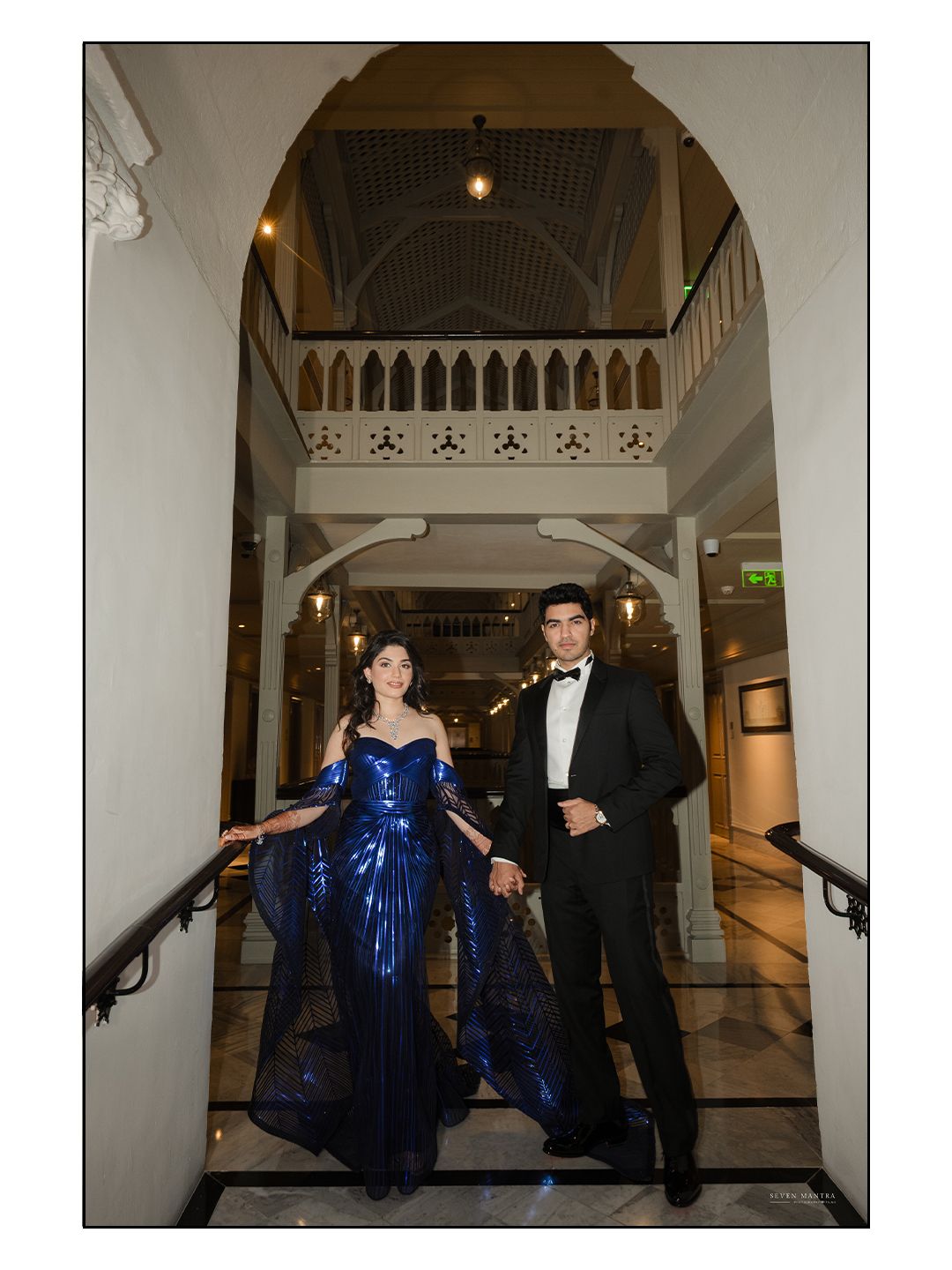 Couple in royal blue gown and black tuxedo pose on grand staircase at luxury wedding venue.