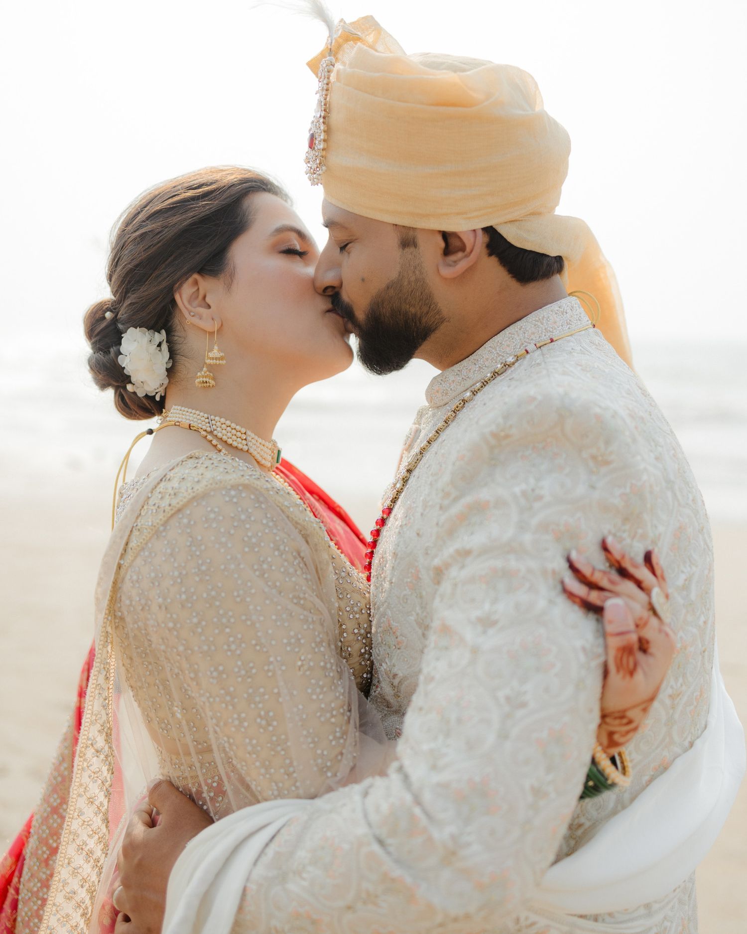 Indian wedding couple sharing a kiss on the beach, bride in ivory outfit, groom in white sherwani with turban.