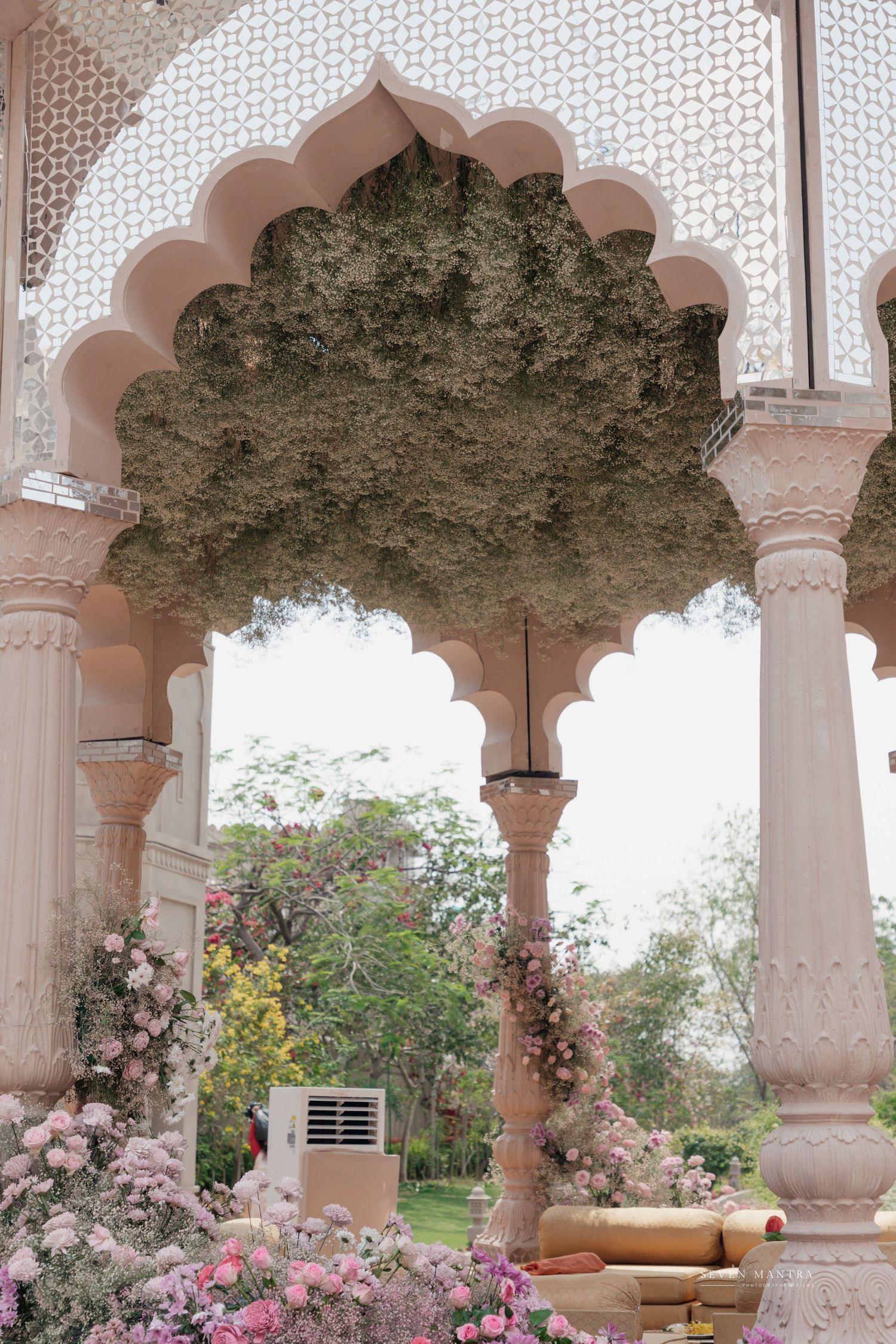 Elegant Indian wedding venue with pink arched pillars, floral decorations and lush green canopy.
