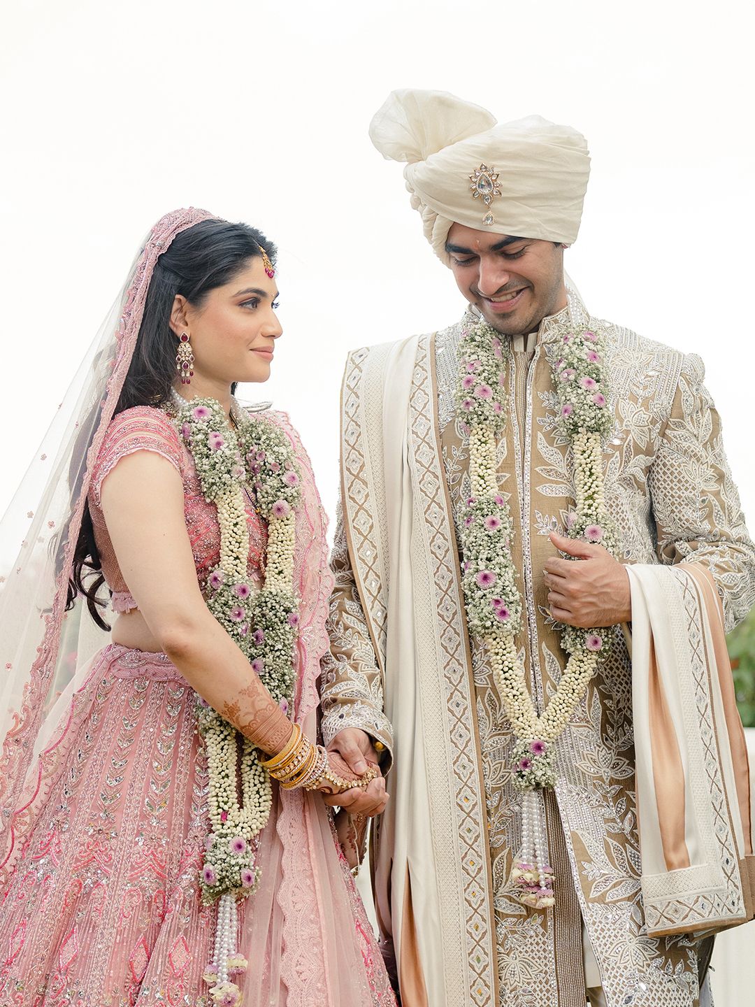 Indian bride in pink lehenga and groom in cream sherwani exchanging garlands at traditional wedding ceremony.