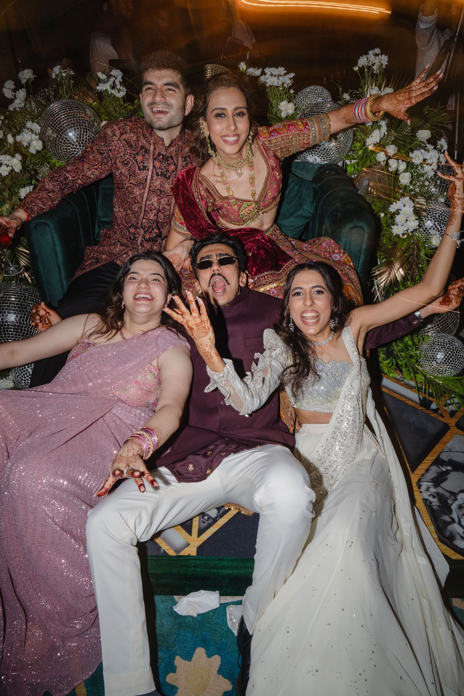 Joyful Indian wedding couple with friends posing playfully on decorated sofa at festive reception.