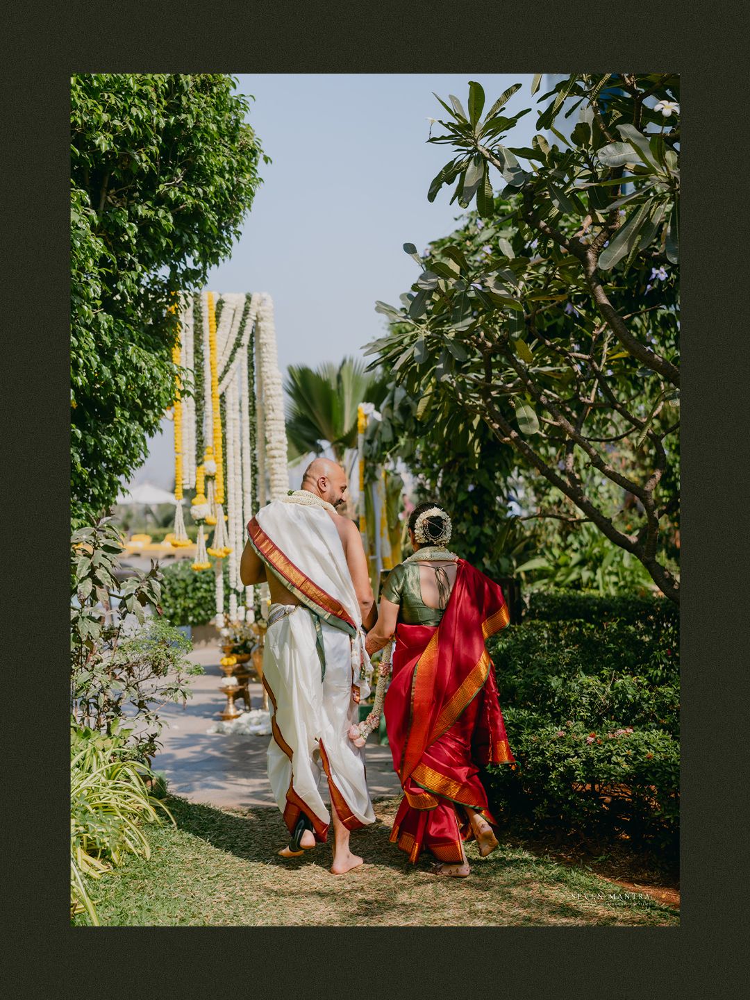 Newlywed couple walks hand in hand along garden path decorated with marigold garlands after South Indian wedding.