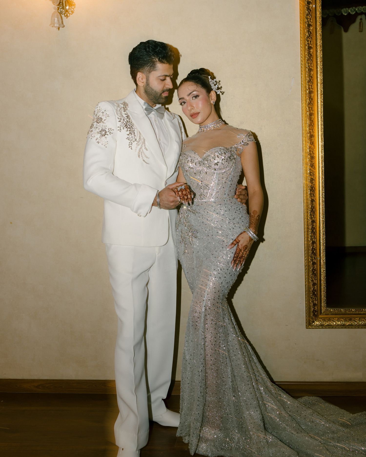 Indian couple posing elegantly, groom in white suit and bride in silver embellished mermaid gown.