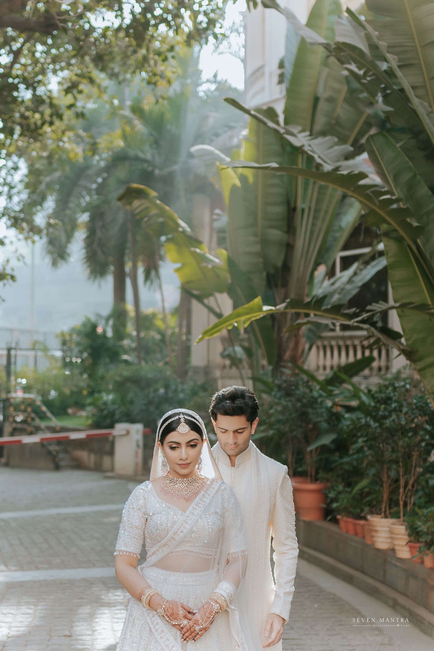 Indian bride and groom in white wedding attire walking together in a lush garden setting.