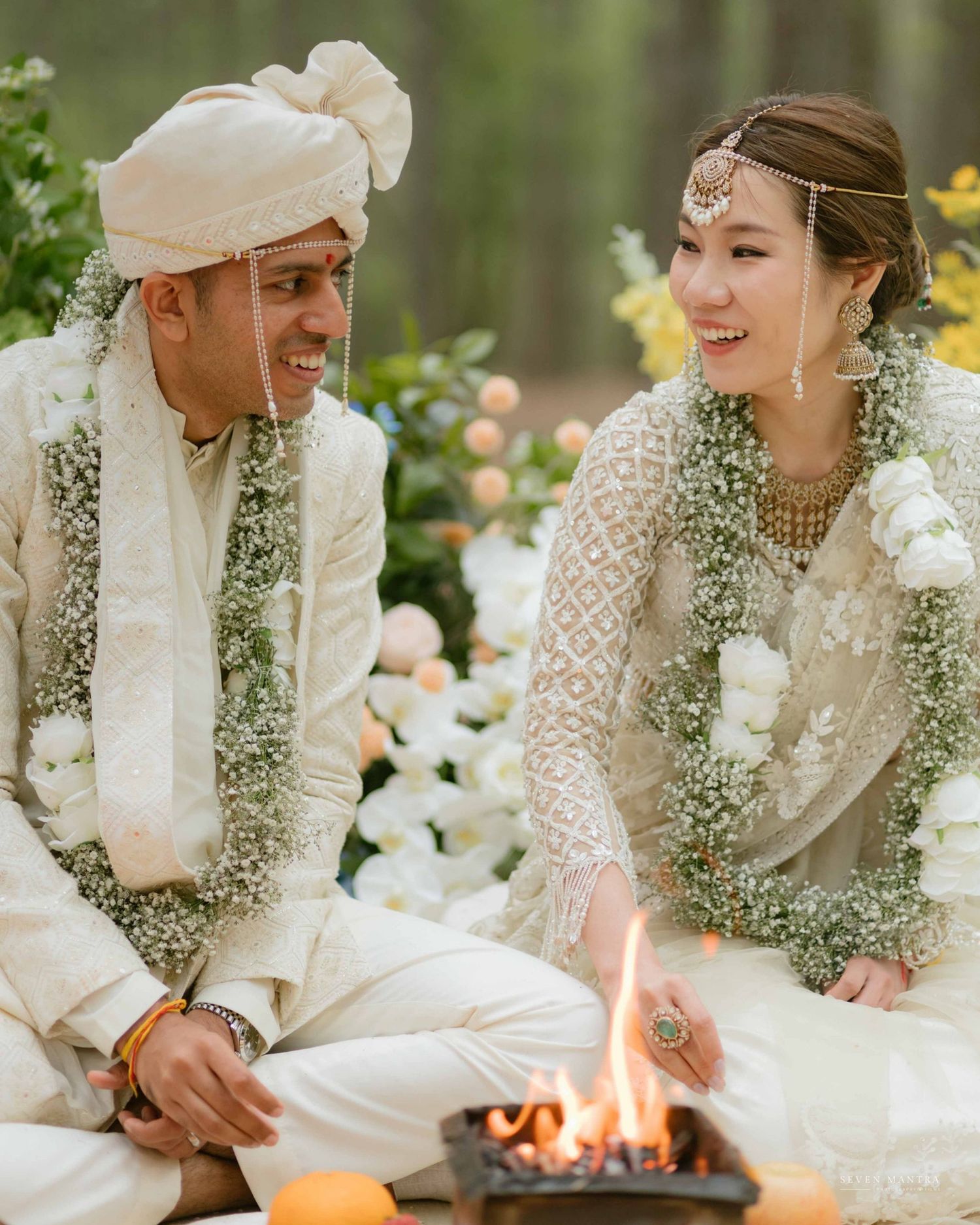 Indian couple in ivory wedding attire smile at each other during a sacred fire ceremony.