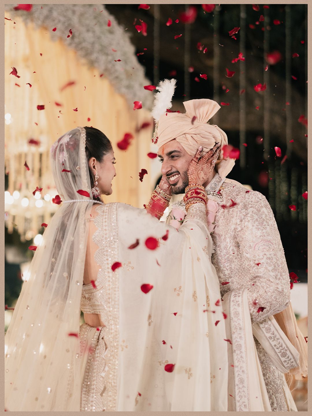 Indian bride and groom in ivory outfits laughing joyfully under red rose petal shower.