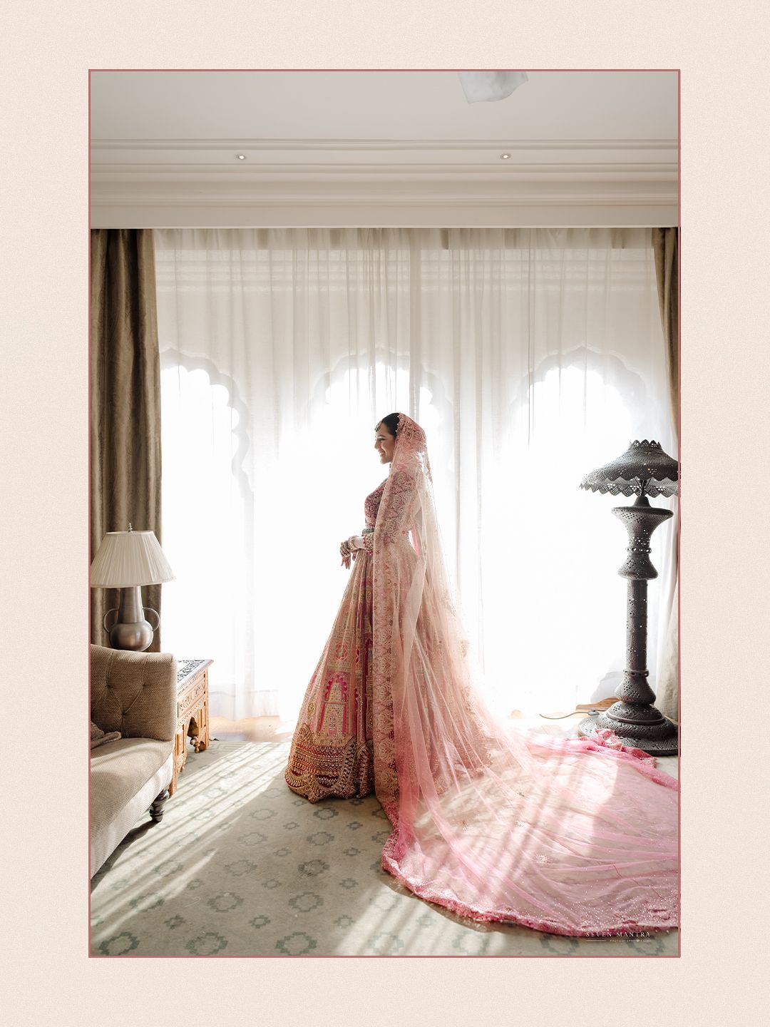 Indian bride in pink embroidered lehenga with long veil stands by window in elegant hotel room.