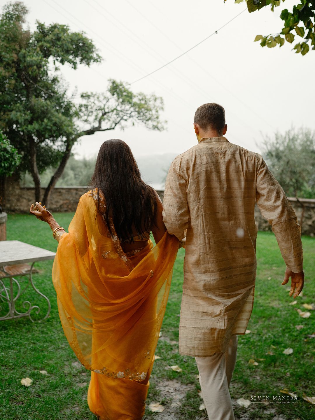 Indian bride in yellow saree and groom in beige kurta walking hand in hand through a lush garden.