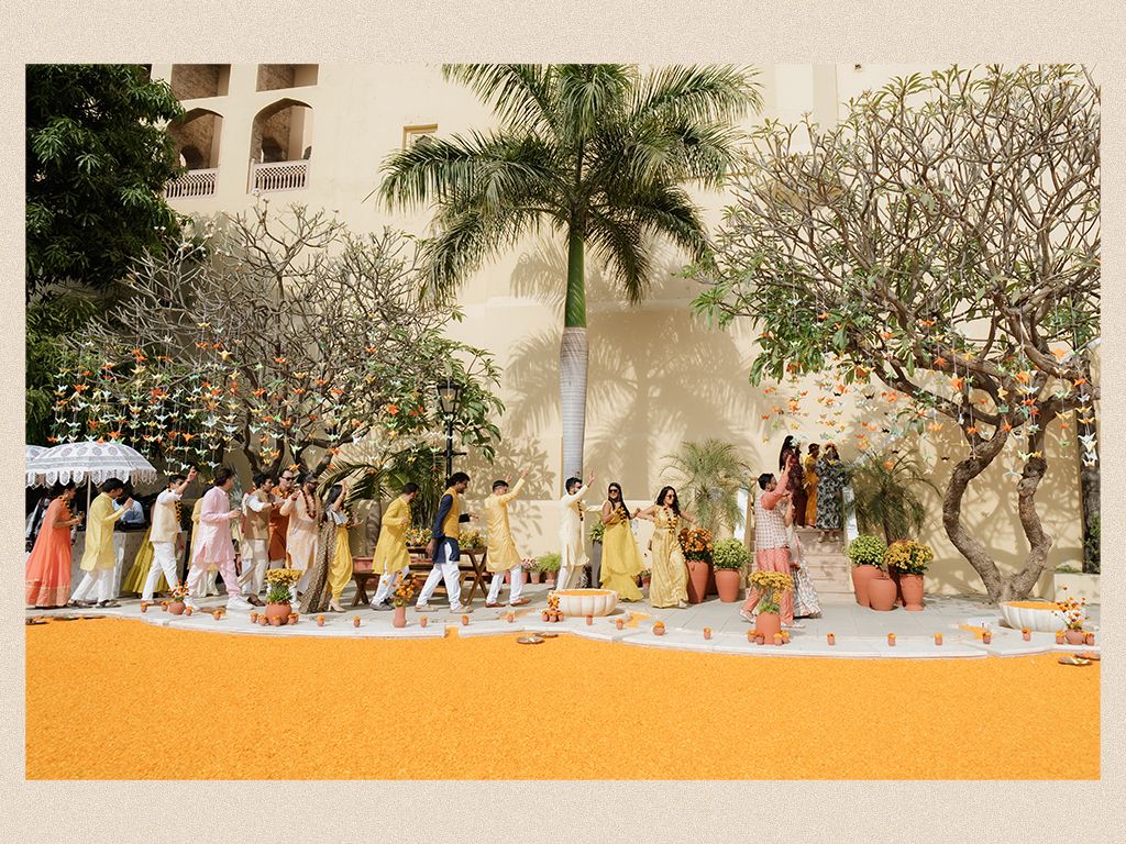 Wedding procession walks along marigold-decorated yellow carpet at elegant outdoor Indian palace venue.