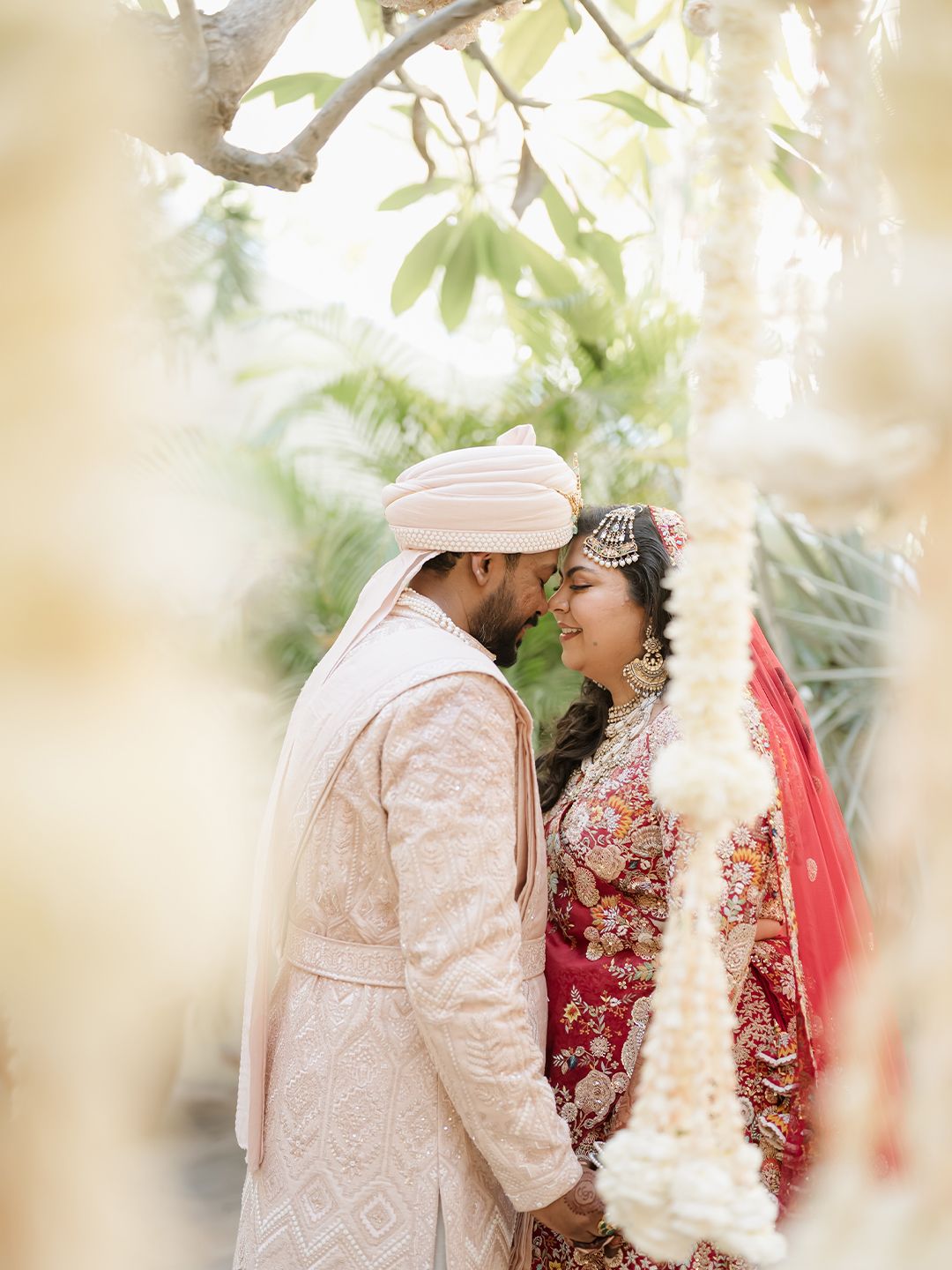 Indian bride and groom share romantic moment in traditional wedding attire, bride in red lehenga, groom in ivory sherwani.