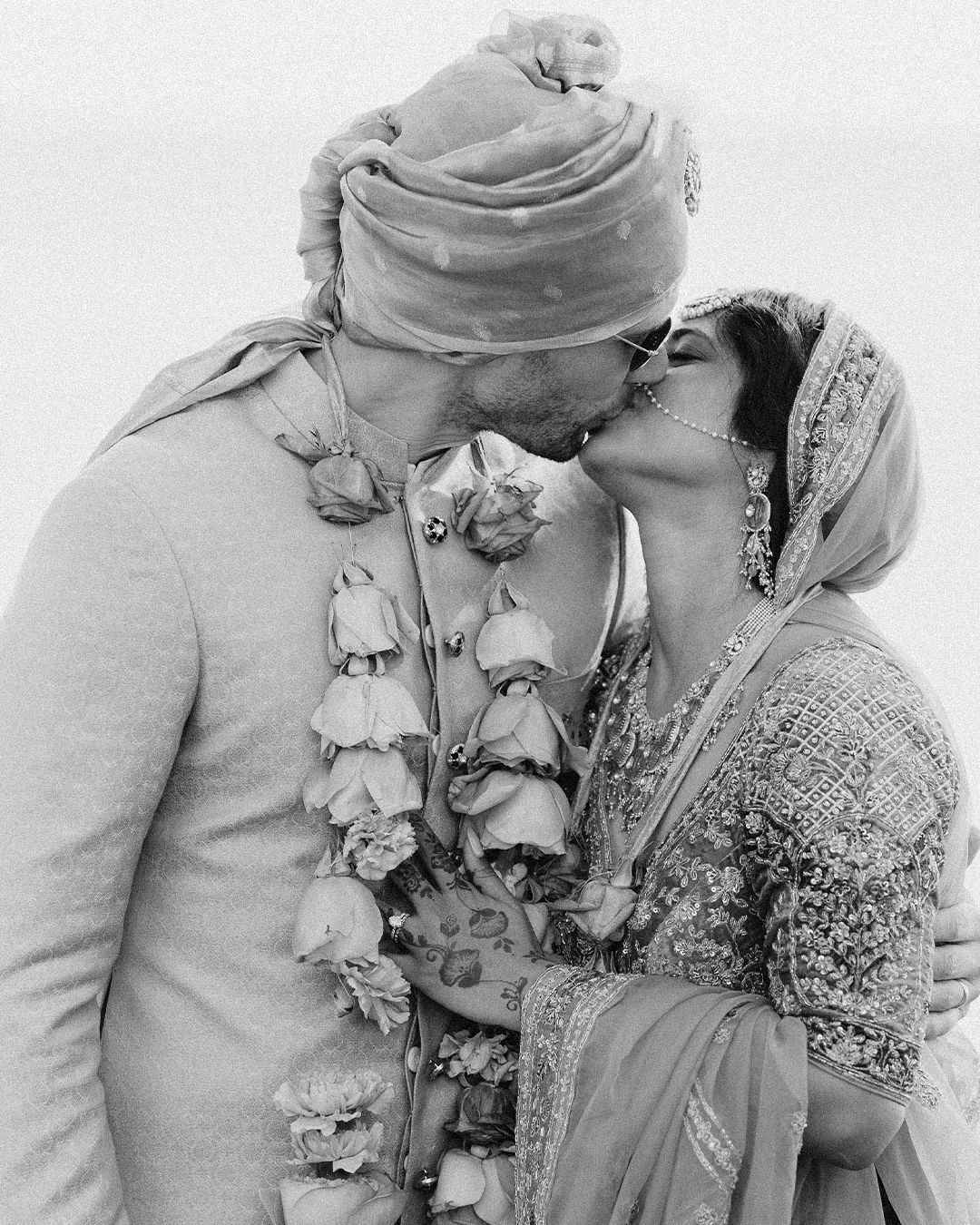Black and white photo of Indian couple sharing a kiss during traditional wedding ceremony with floral garlands.