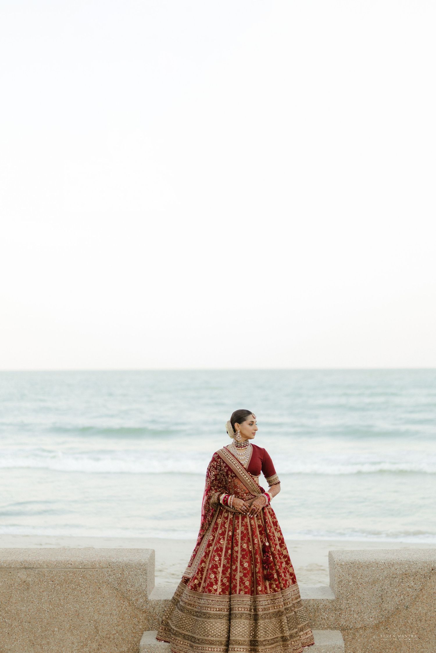 Indian bride in red embroidered lehenga standing by ocean at beach wedding venue.