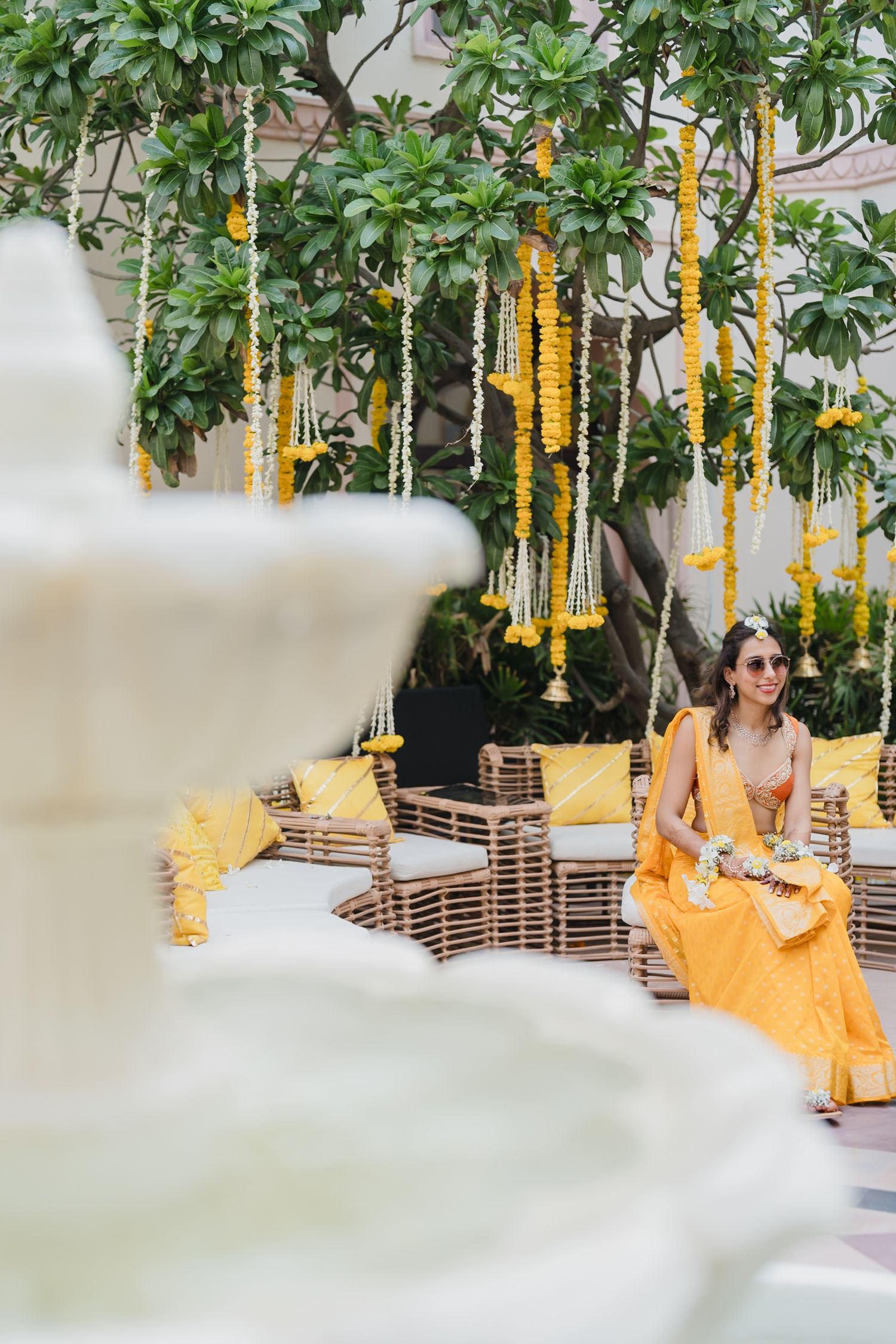 Woman in yellow lehenga sits among rattan furniture decorated with marigold floral hangings at Indian wedding.