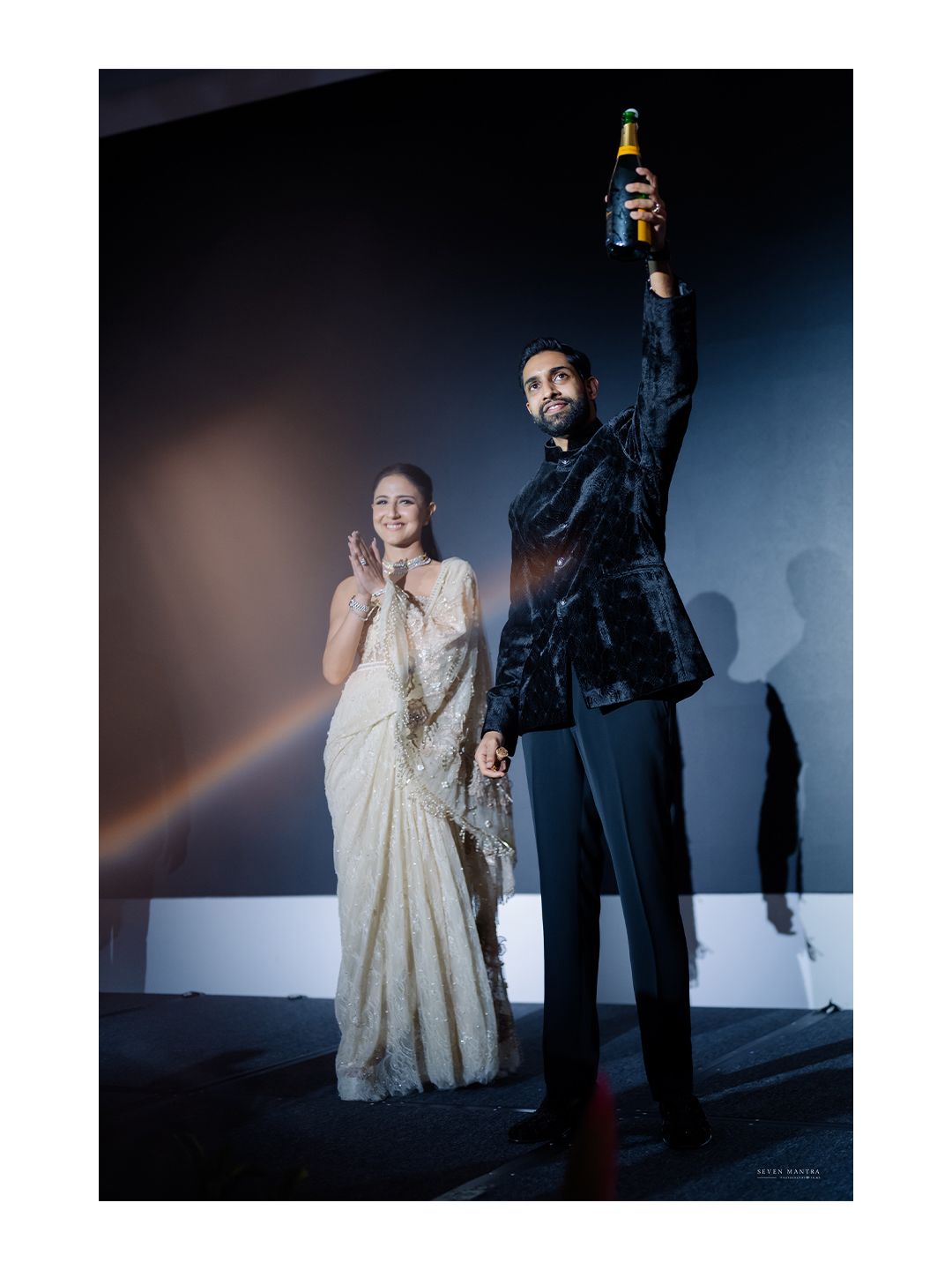 Indian groom in black velvet sherwani raising champagne bottle while bride in white saree claps at wedding.