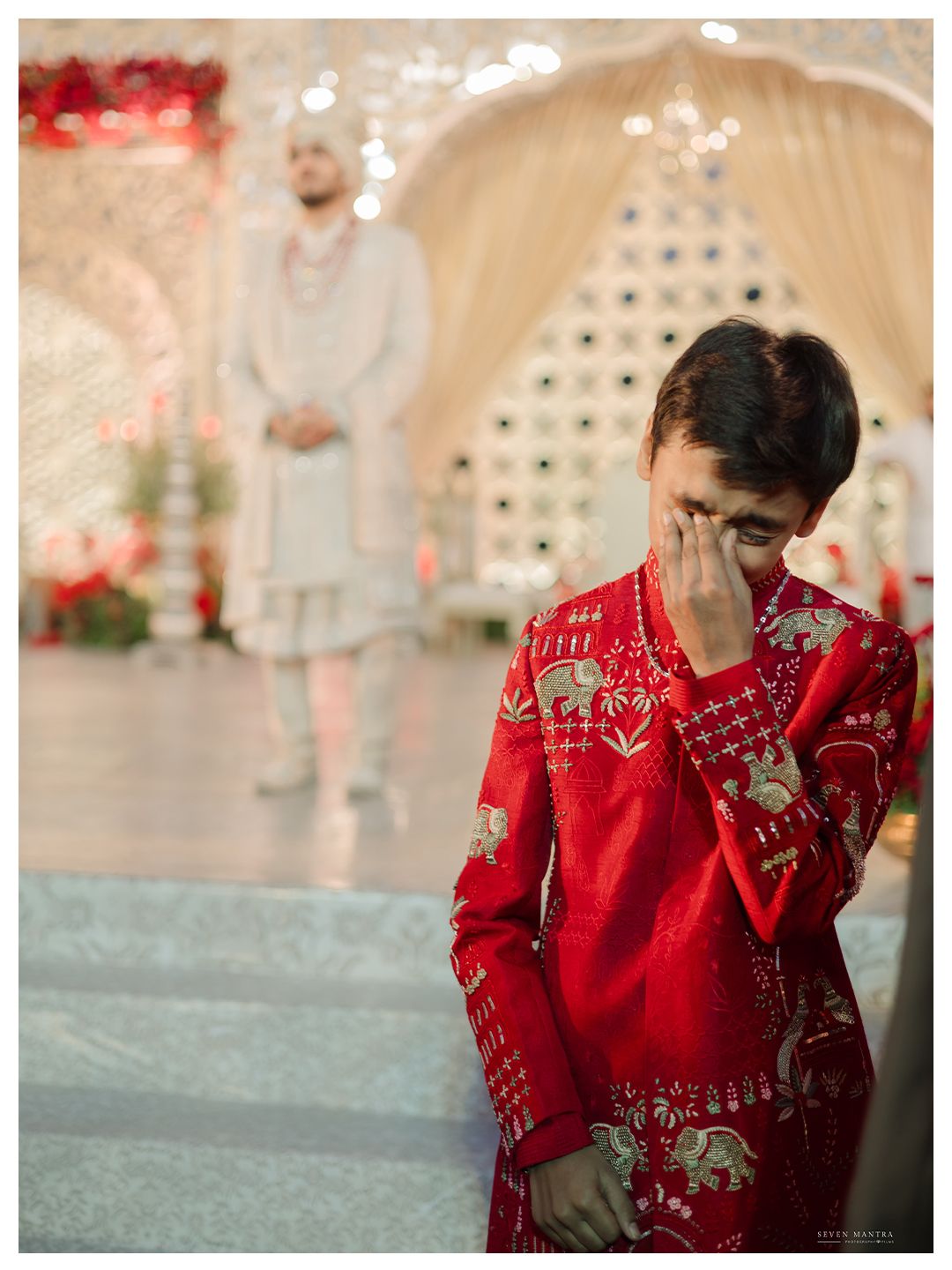 Emotional young boy in red embroidered sherwani wiping tears at Indian wedding ceremony.
