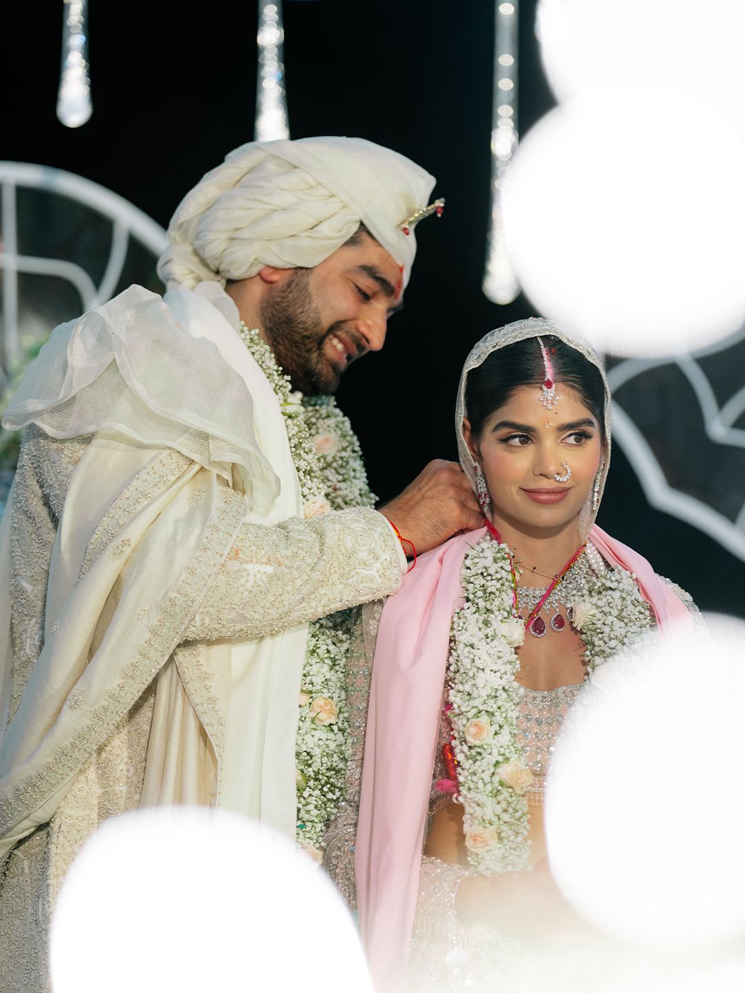 Indian groom in white sherwani and turban placing garland on bride during traditional wedding ceremony.