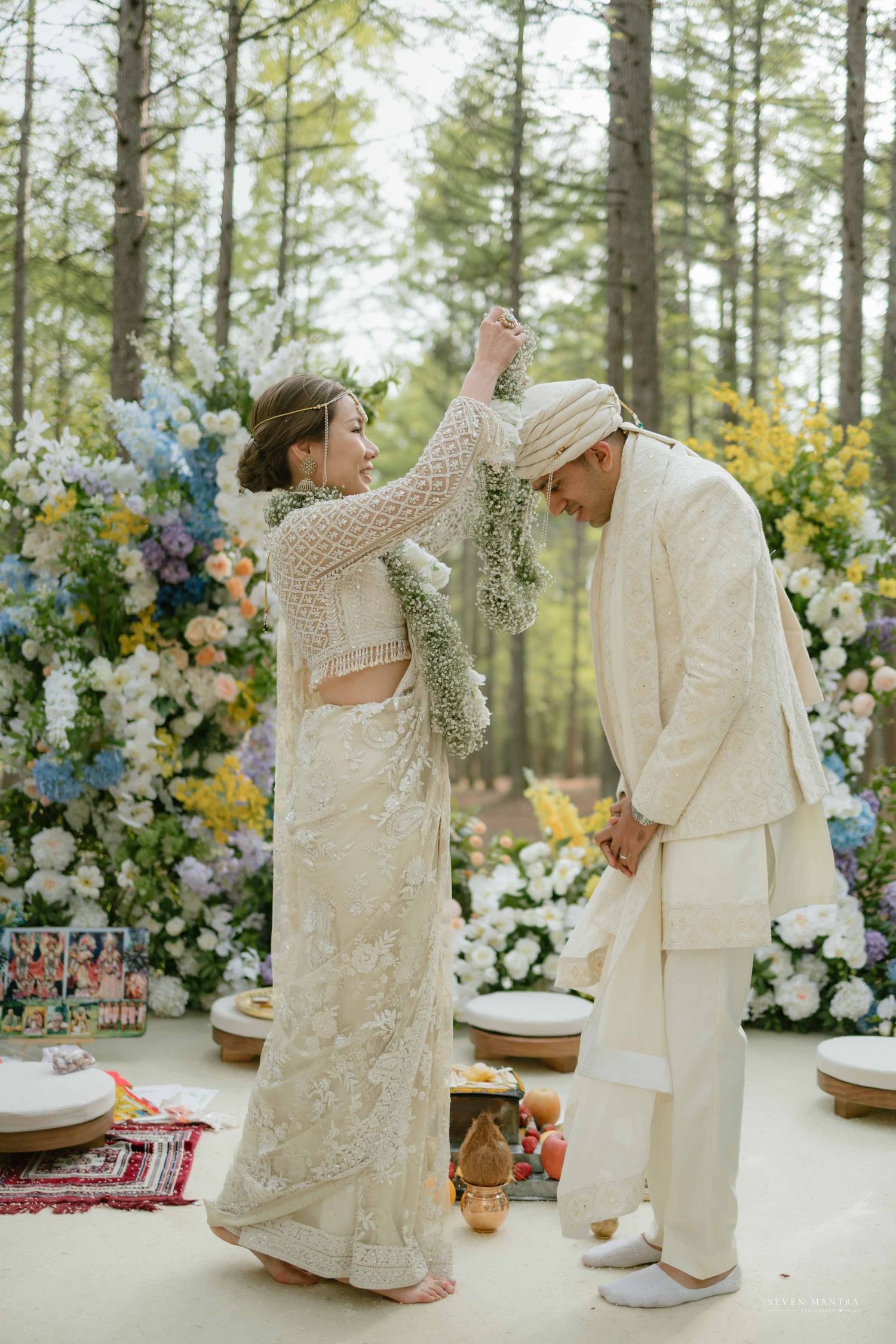 Bride places floral garland on groom during outdoor Hindu wedding ceremony in forest setting.