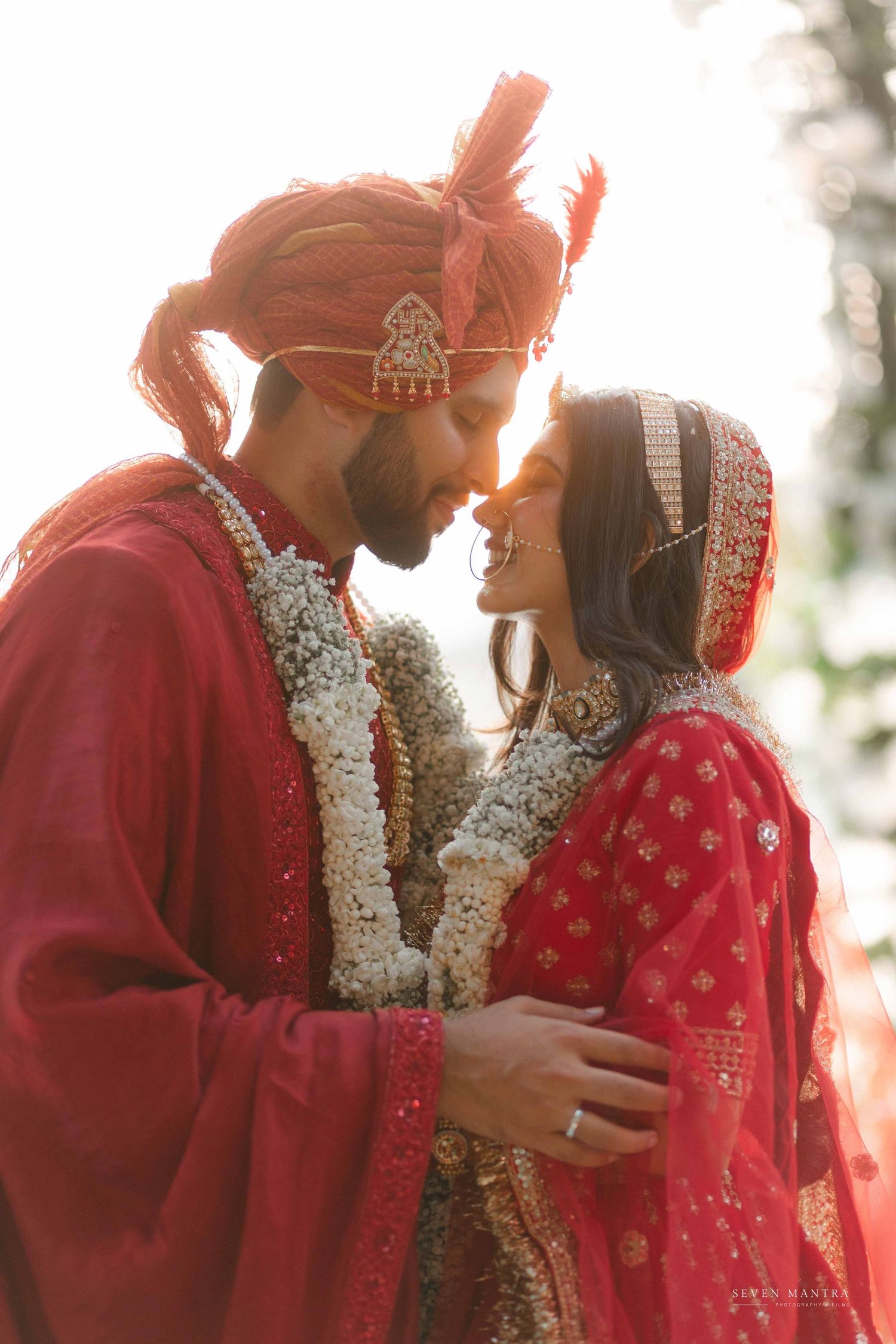 Indian couple in traditional red wedding attire sharing romantic moment in golden sunset light.